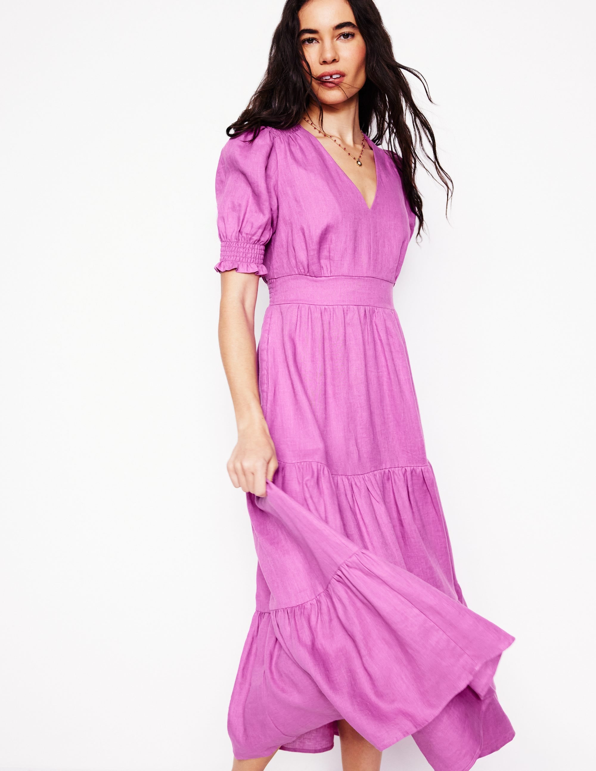A woman wearing a purple linen midi dress with puff sleeves and a tiered skirt, standing against a plain background.