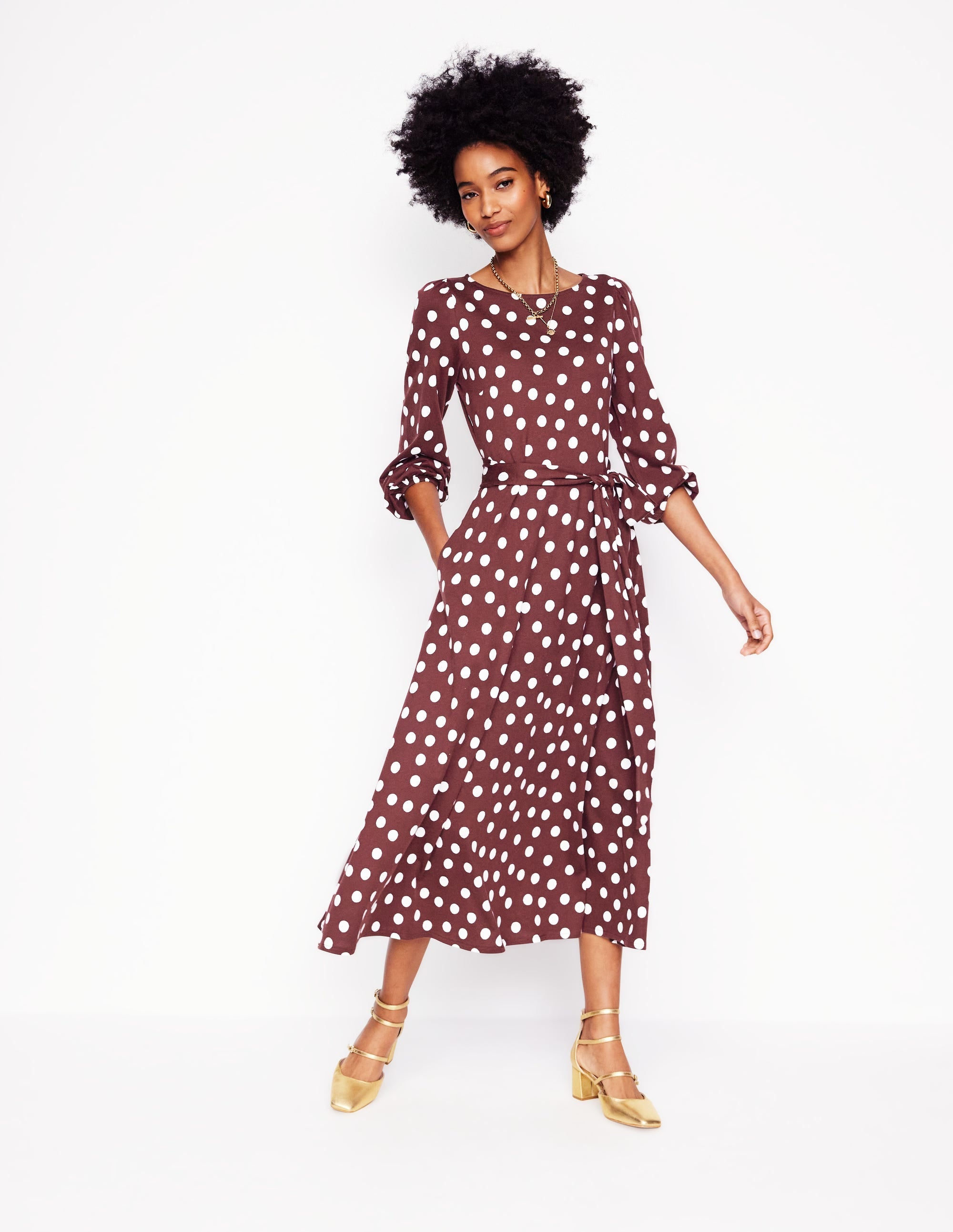 A woman wearing a chestnut brown midi dress with white abstract dots, long sleeves with gathered cuffs, and a self-fabric belt, standing against a white background.