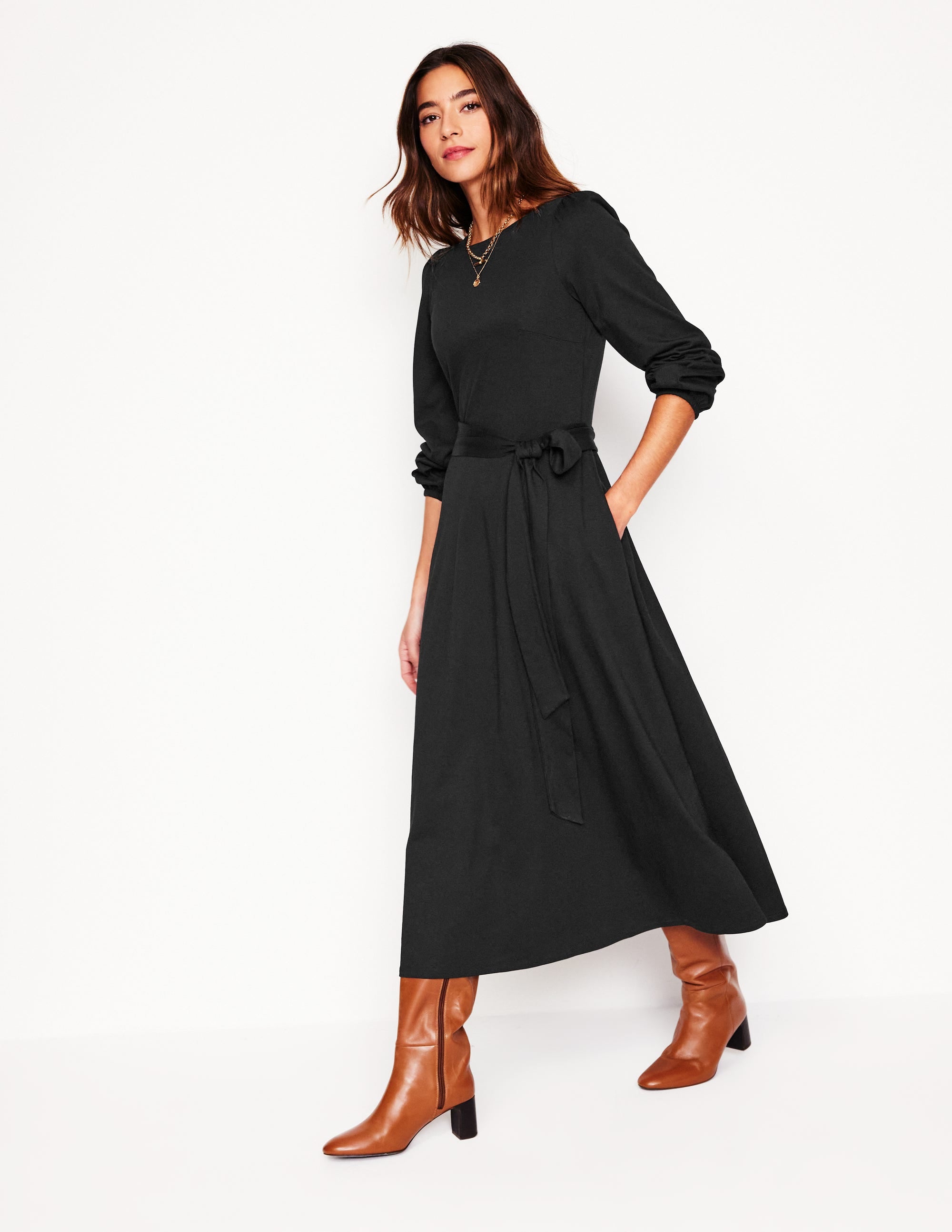 A woman wearing a black midi dress with long sleeves, a self-fabric belt, and side pockets, paired with brown knee-high boots.