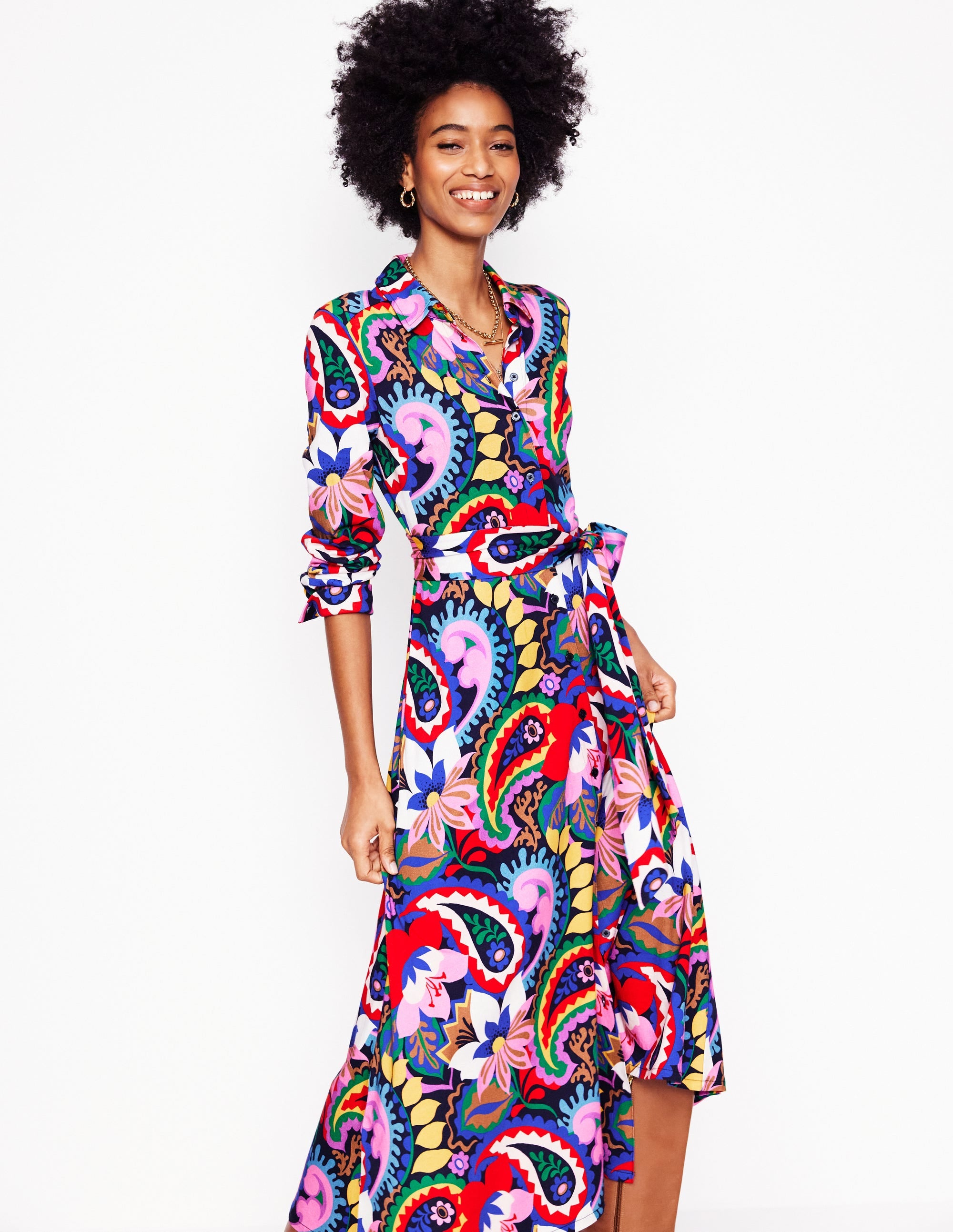 A woman wearing a colorful, long-sleeved midi shirt dress with a paisley and floral pattern, a waist belt, and side pockets.