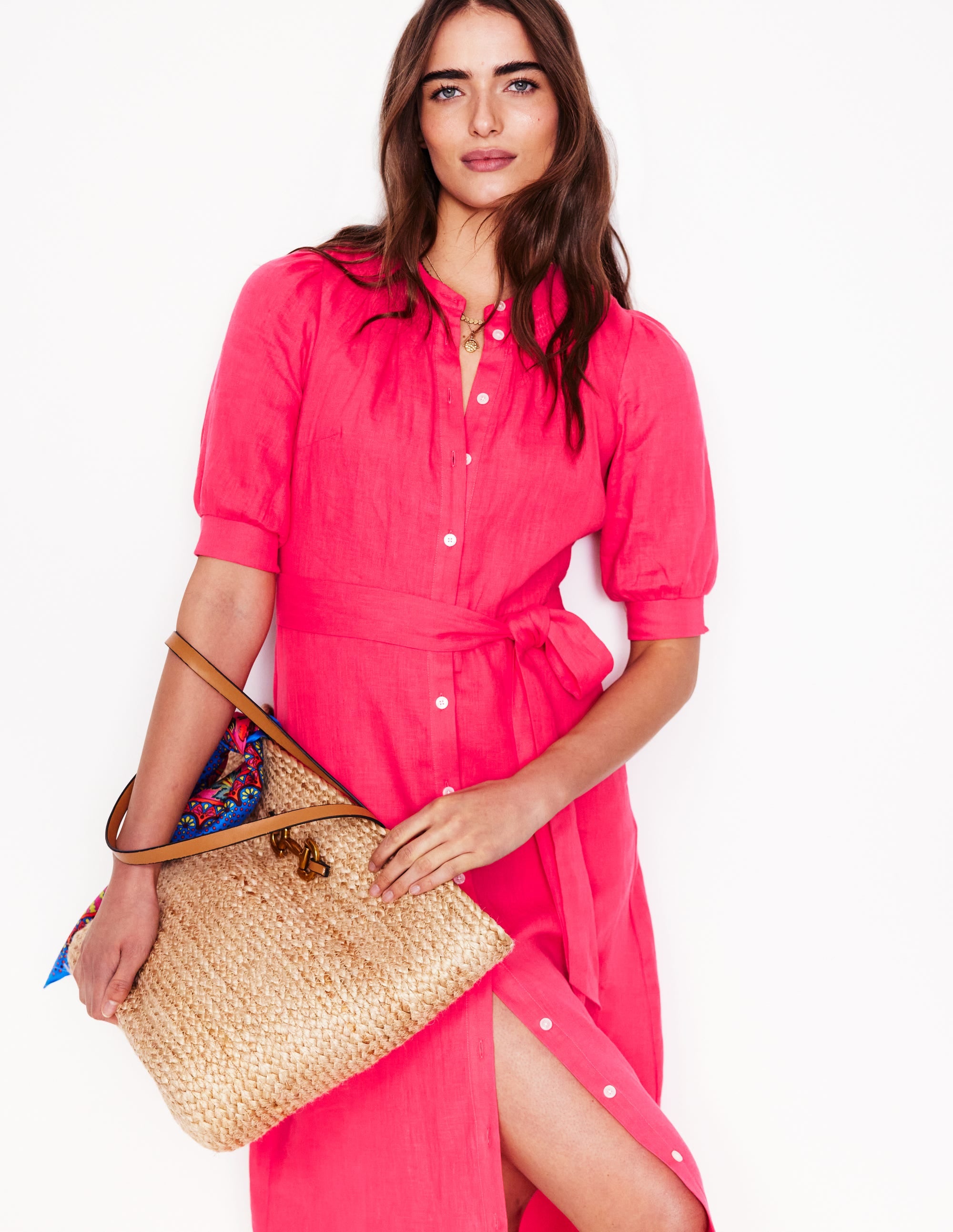 A woman wearing a pink linen midi dress with gathered sleeves, a smocked neckline, and a front button closure, holding a woven bag.