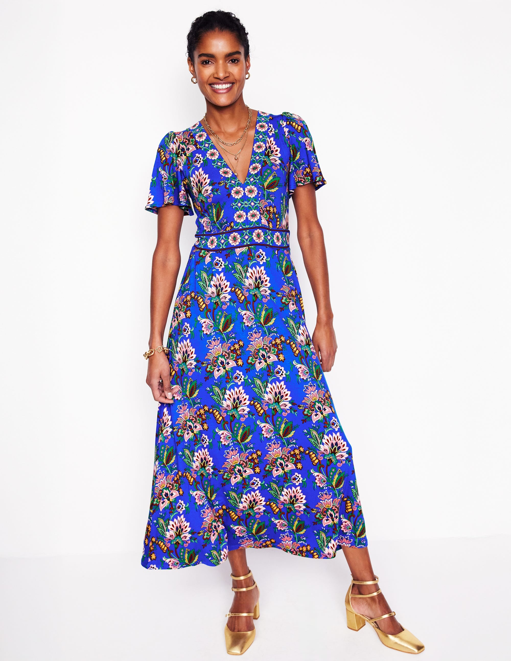 A woman wearing a blue floral maxi dress with short flutter sleeves, V-neckline, and side seam pockets, standing against a white background.