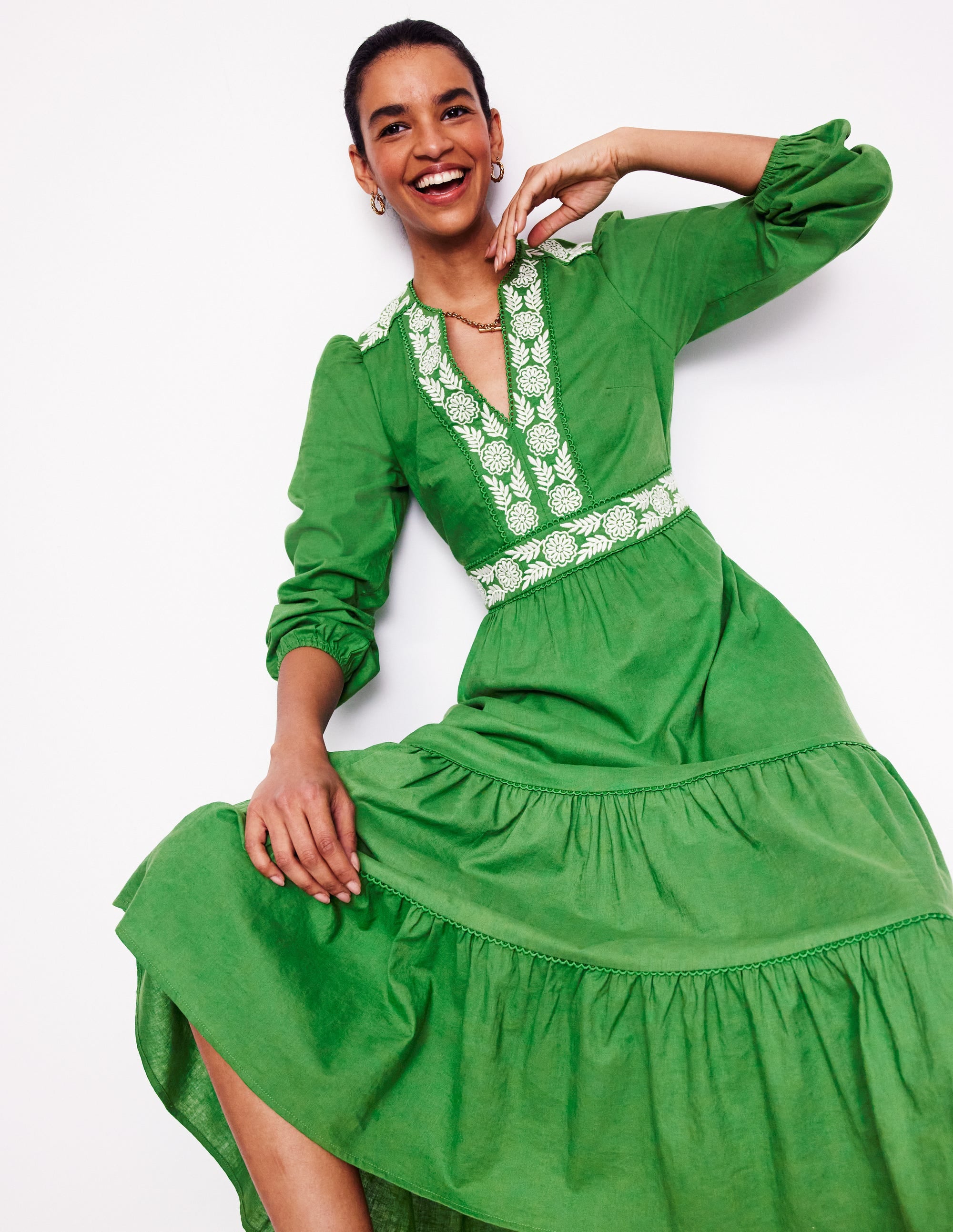 A woman wearing a bright green linen blend midi dress with embroidered white floral details, long sleeves, and a flared skirt.
