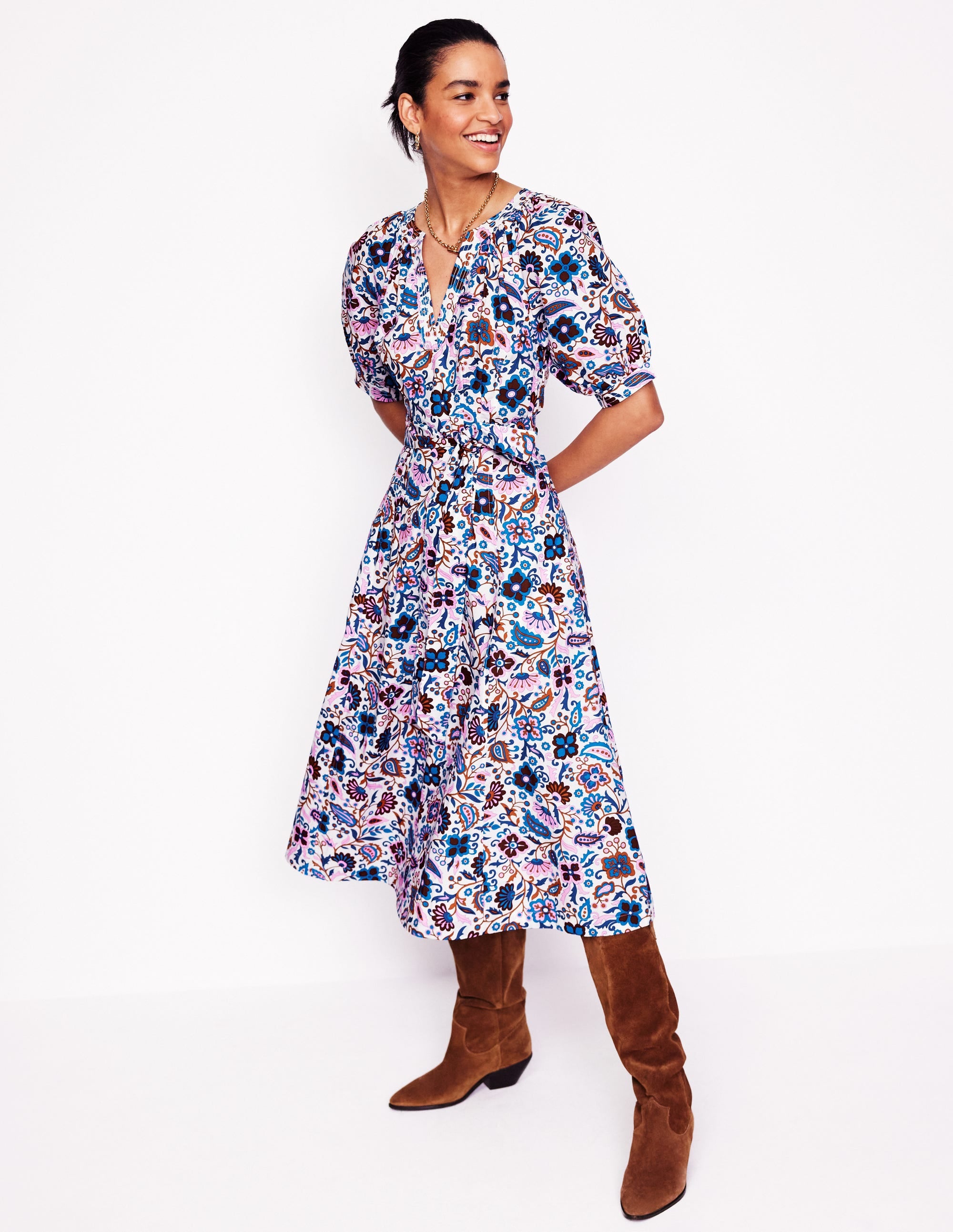 A woman wearing a multicolour floral and paisley patterned midi dress with puff sleeves, a tie belt at the waist, and a V-neckline, paired with brown knee-high boots.
