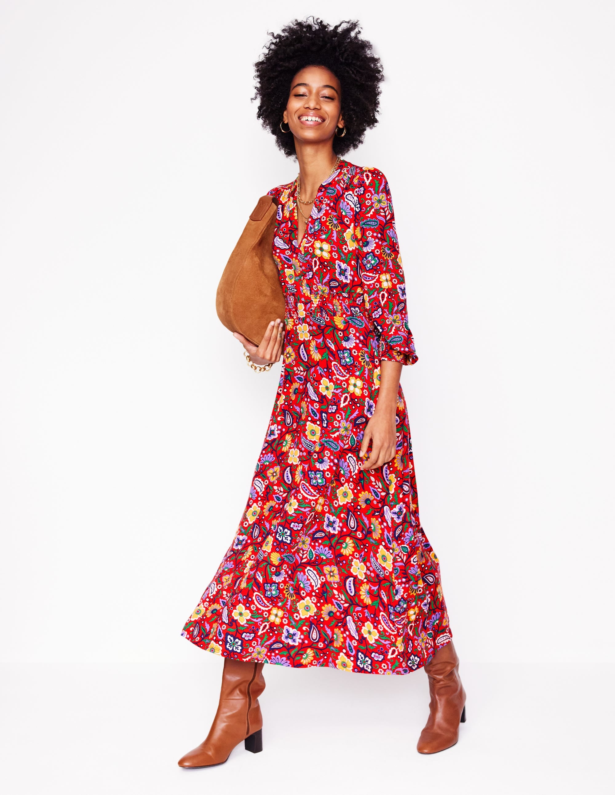 A woman wearing a red floral midi dress with a notch neckline and smocked waist, paired with brown boots and a tan bag.