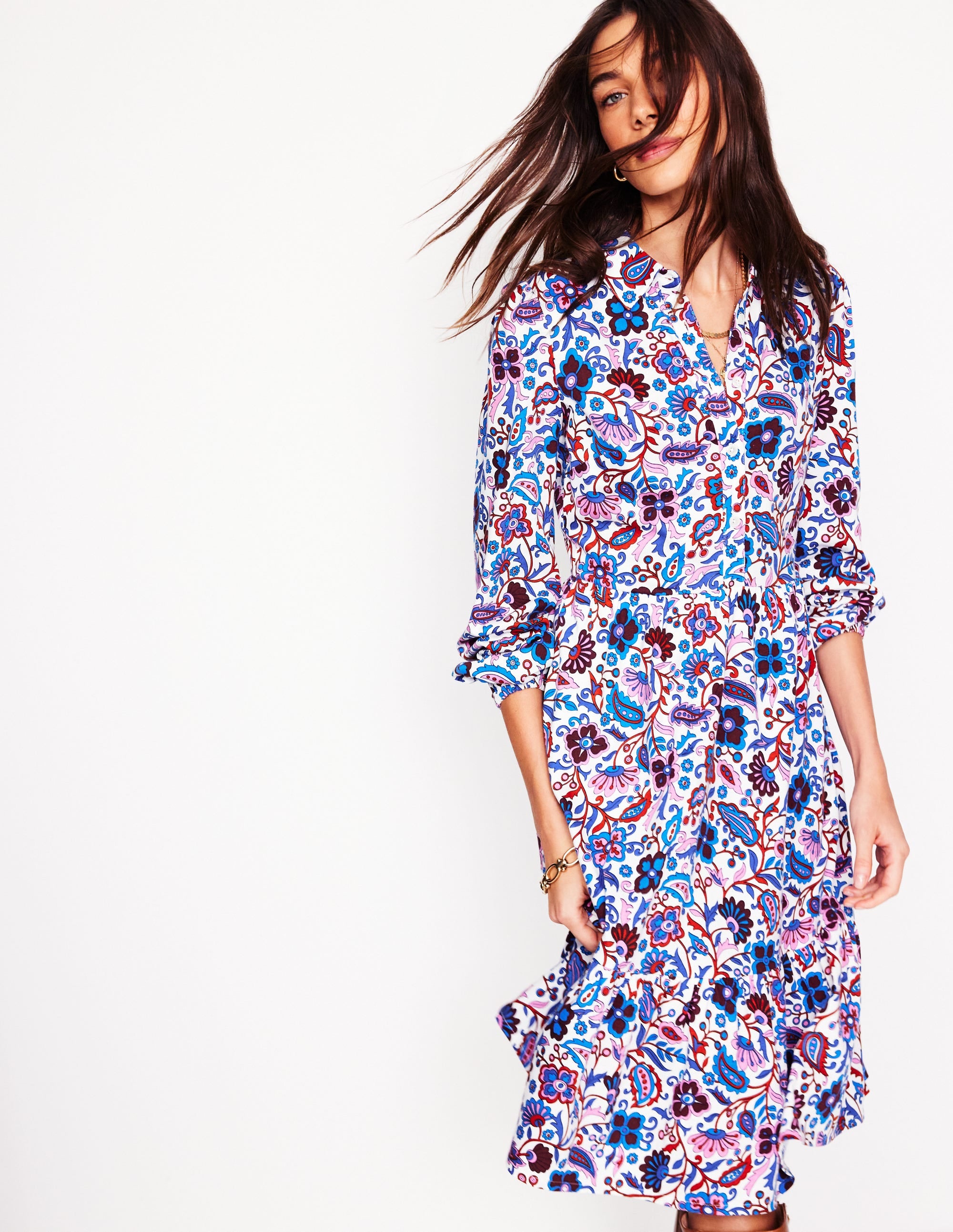 A woman wearing a long-sleeved, tiered jersey dress with a vibrant floral pattern in shades of blue, purple, red, and pink, with a collar and button-down front.