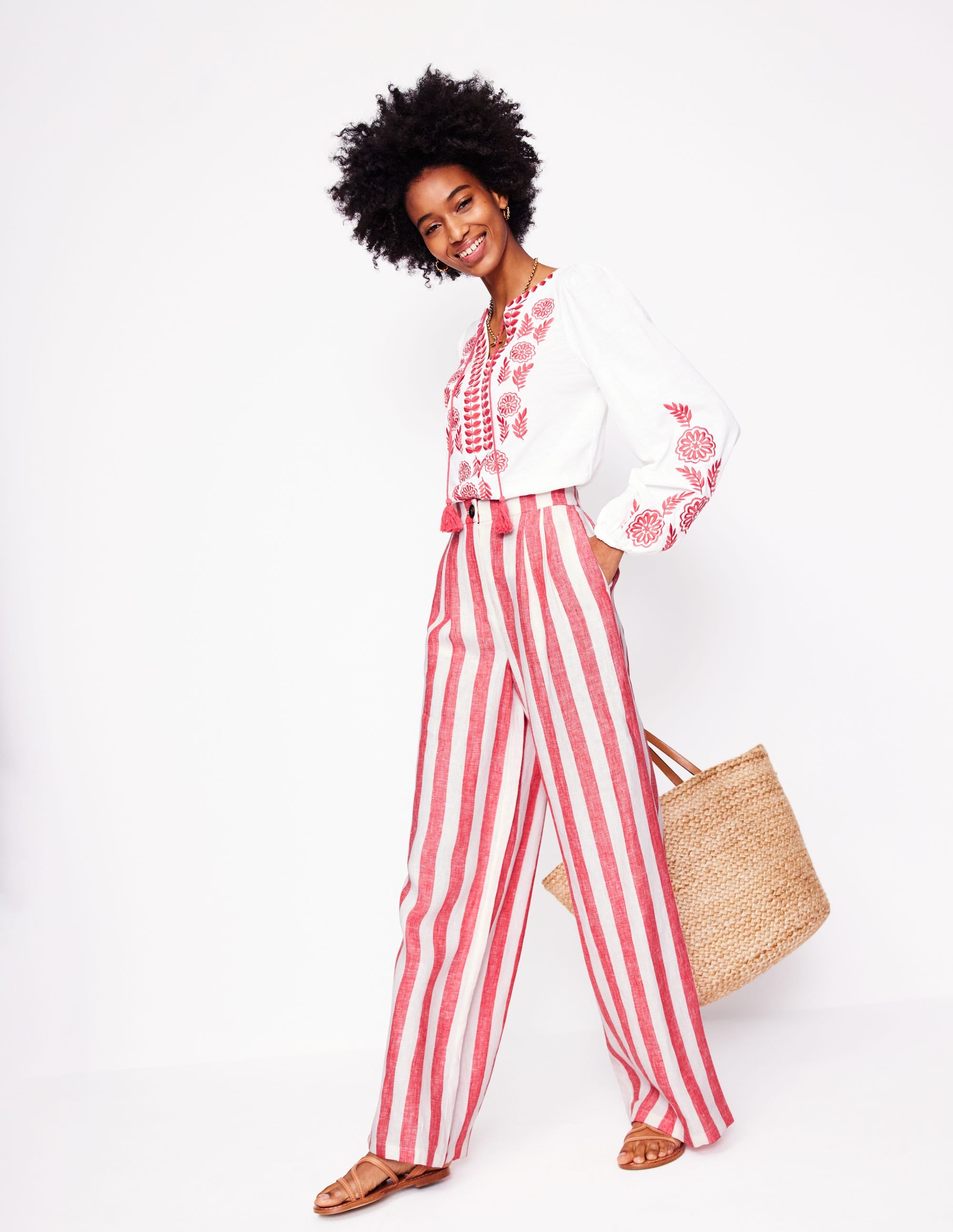 A woman wearing high-waisted, wide-leg linen trousers with red and white stripes, paired with a white embroidered blouse, holding a straw bag.