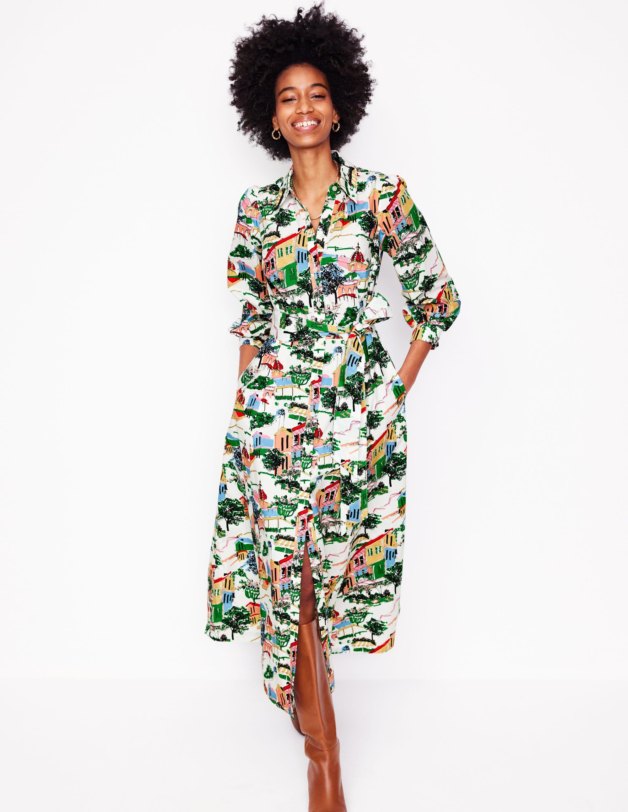 A woman wearing a colorful, painted village print shirt dress with long sleeves, a collar, a button-down front, a waist belt, side pockets, and a maxi length.