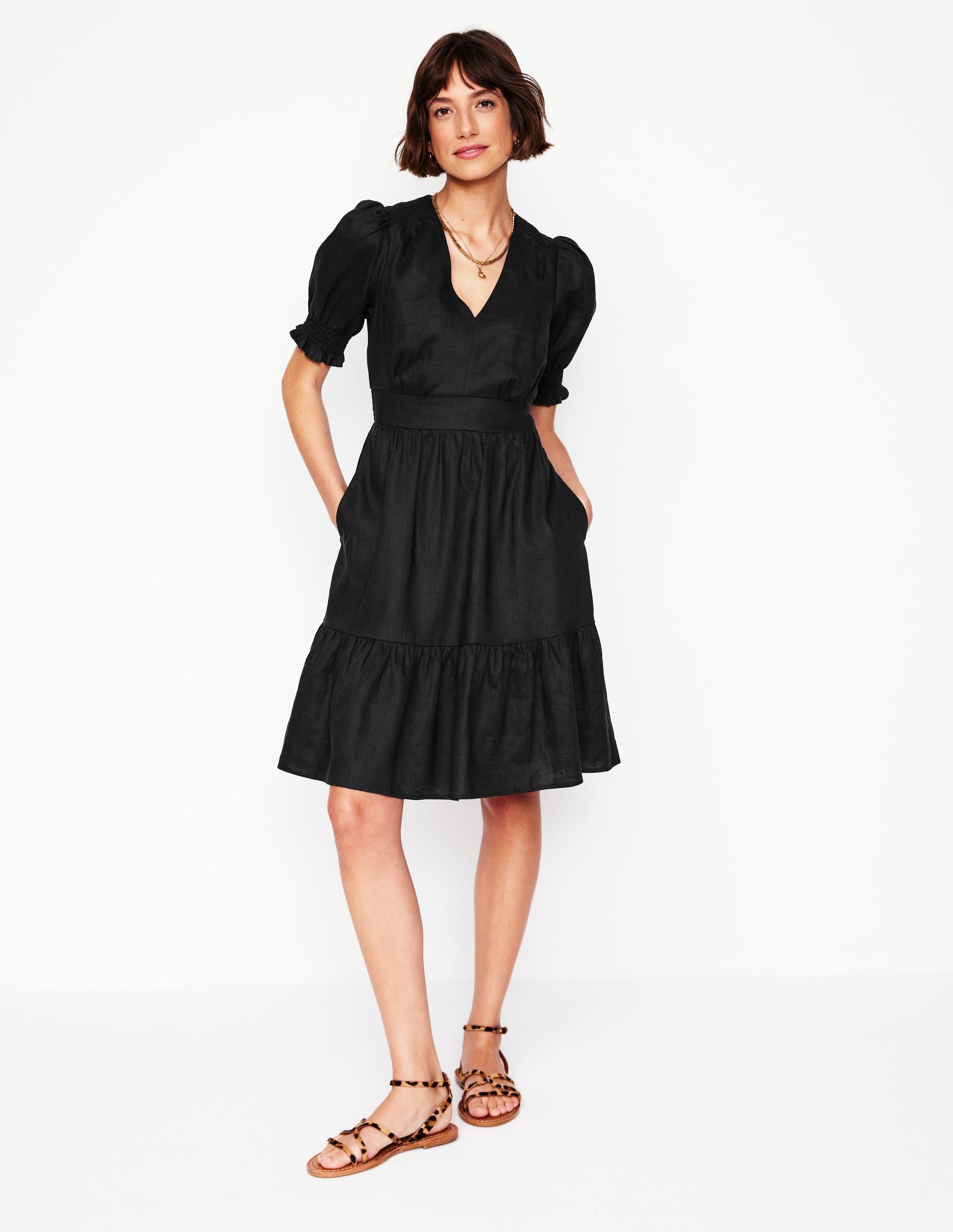 A woman wearing a black linen short dress with puff sleeves, a tiered skirt, and side pockets, standing against a white background.