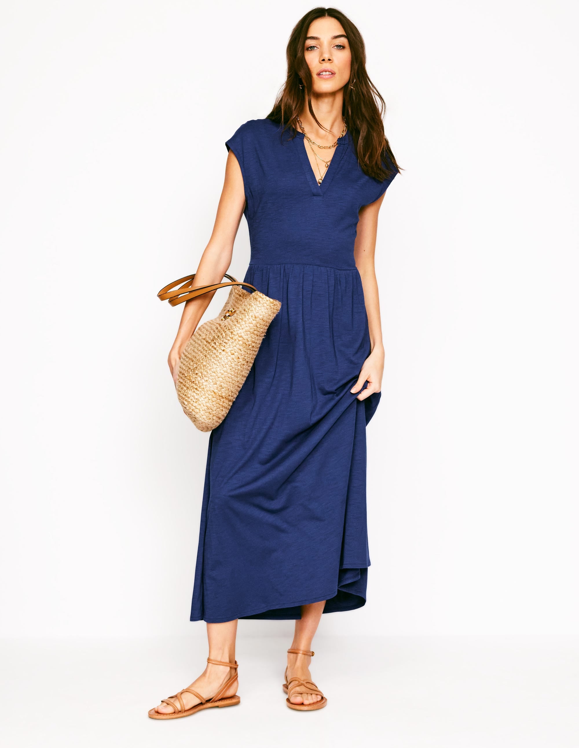 A woman wearing a blue midi dress with a V-neckline, short sleeves, and a flowing skirt, holding a straw bag and wearing sandals.