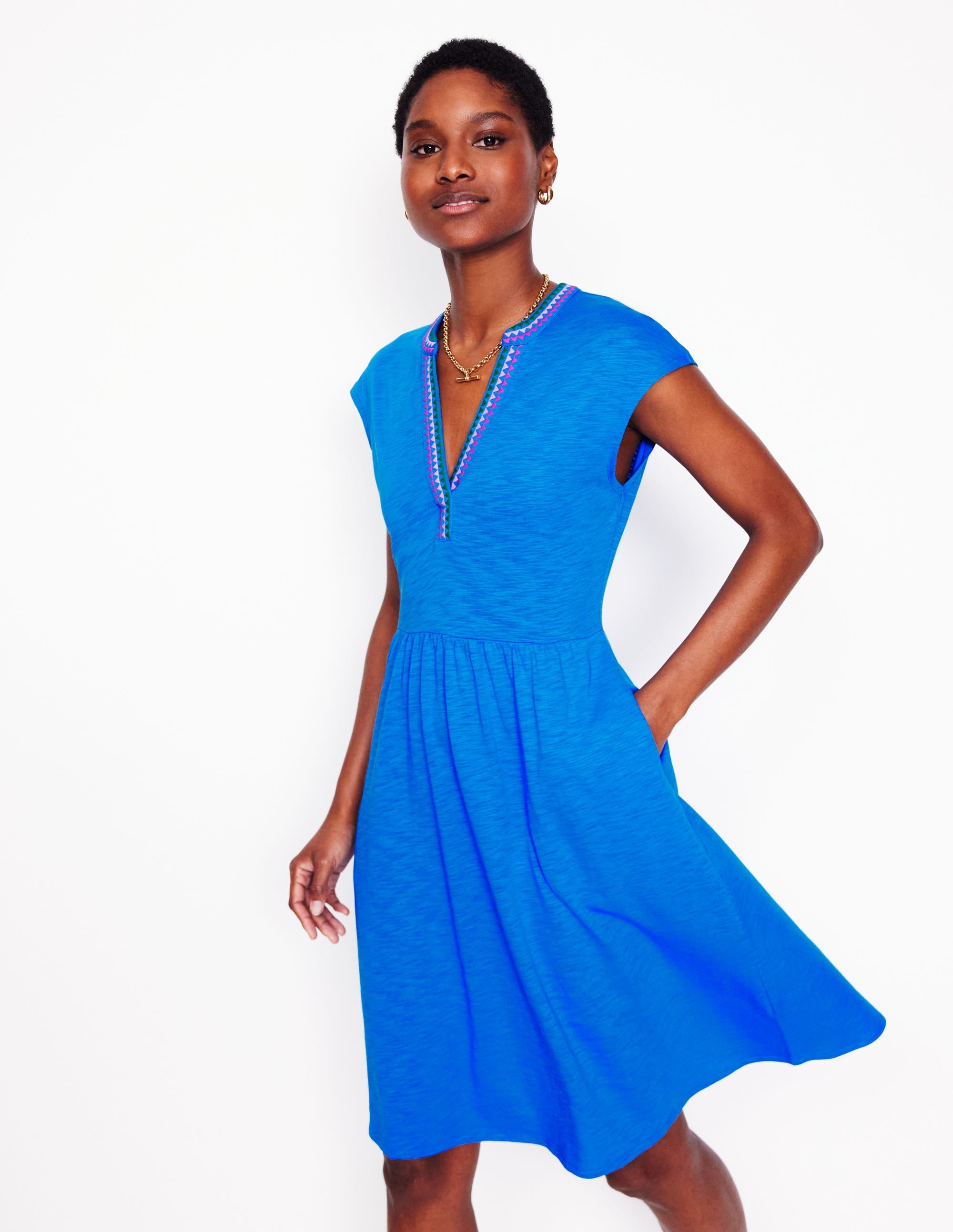 A woman wearing a bright blue embroidered short dress with a V-neckline and sleeveless design, standing against a white background.