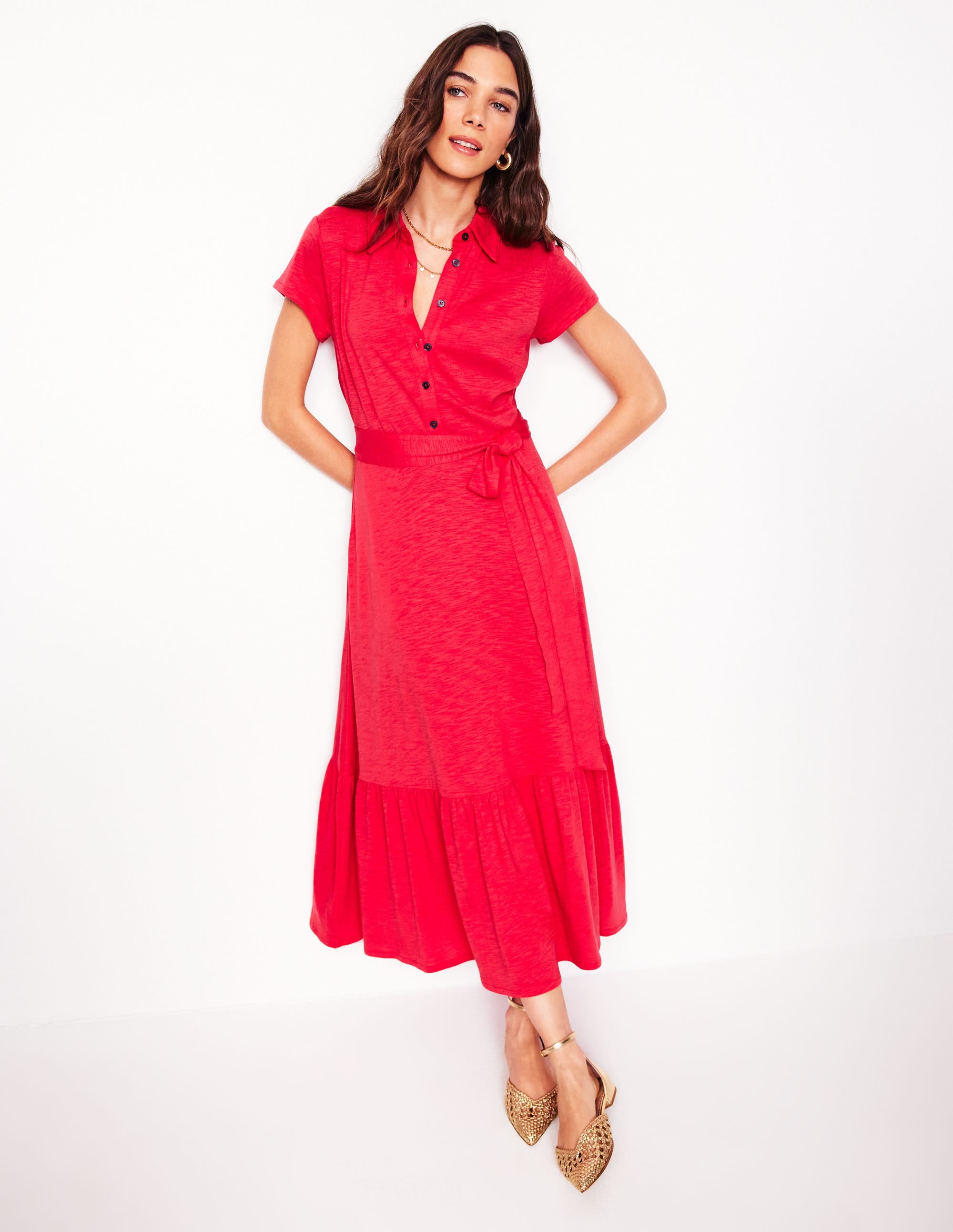 A woman wearing a jewel red jersey shirt dress with a collar, cap sleeves, button-down front, tiered skirt, and side pockets.