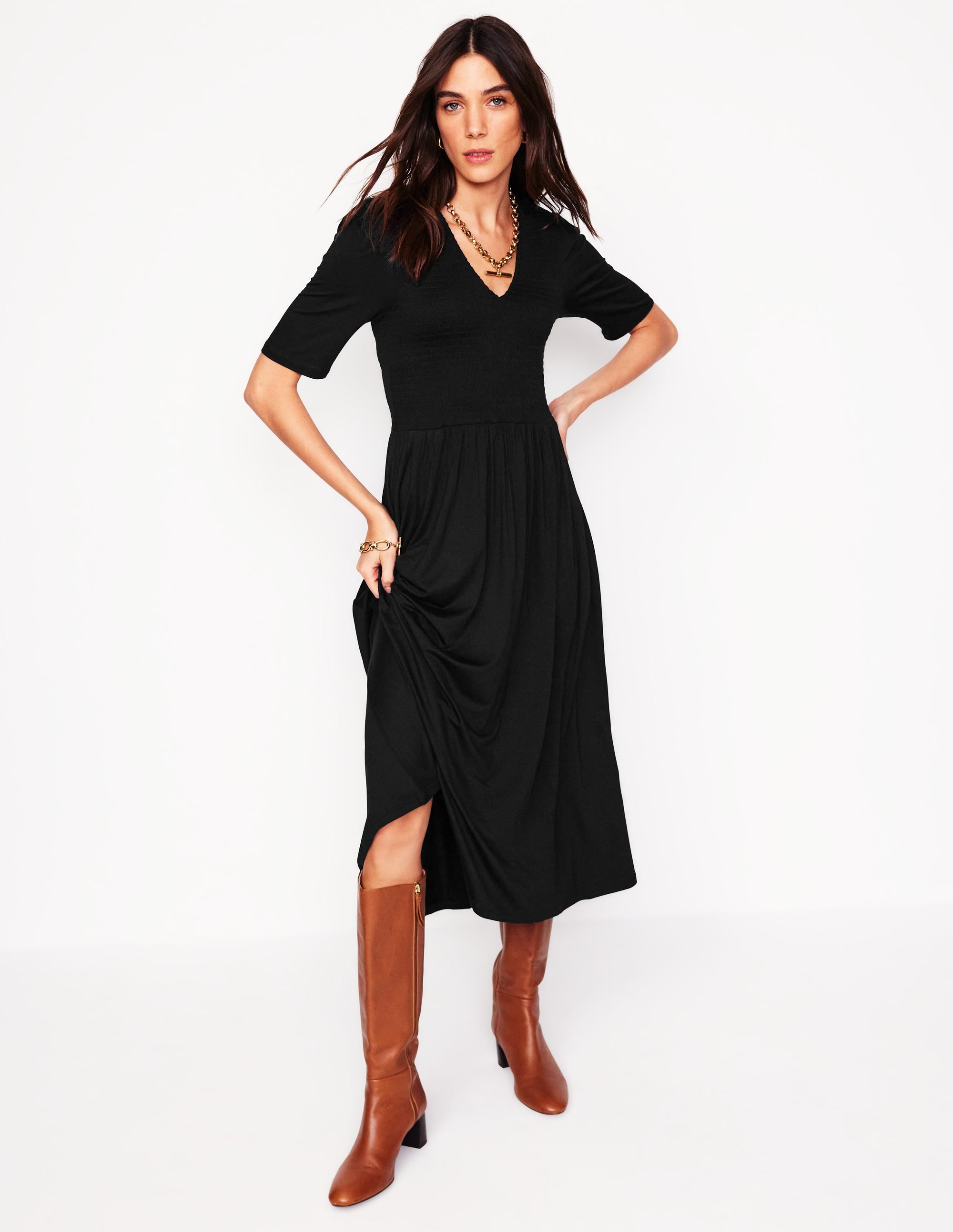 A woman wearing a black smocked jersey dress with a V-neck, short sleeves, and a flowy skirt, paired with brown knee-high boots.