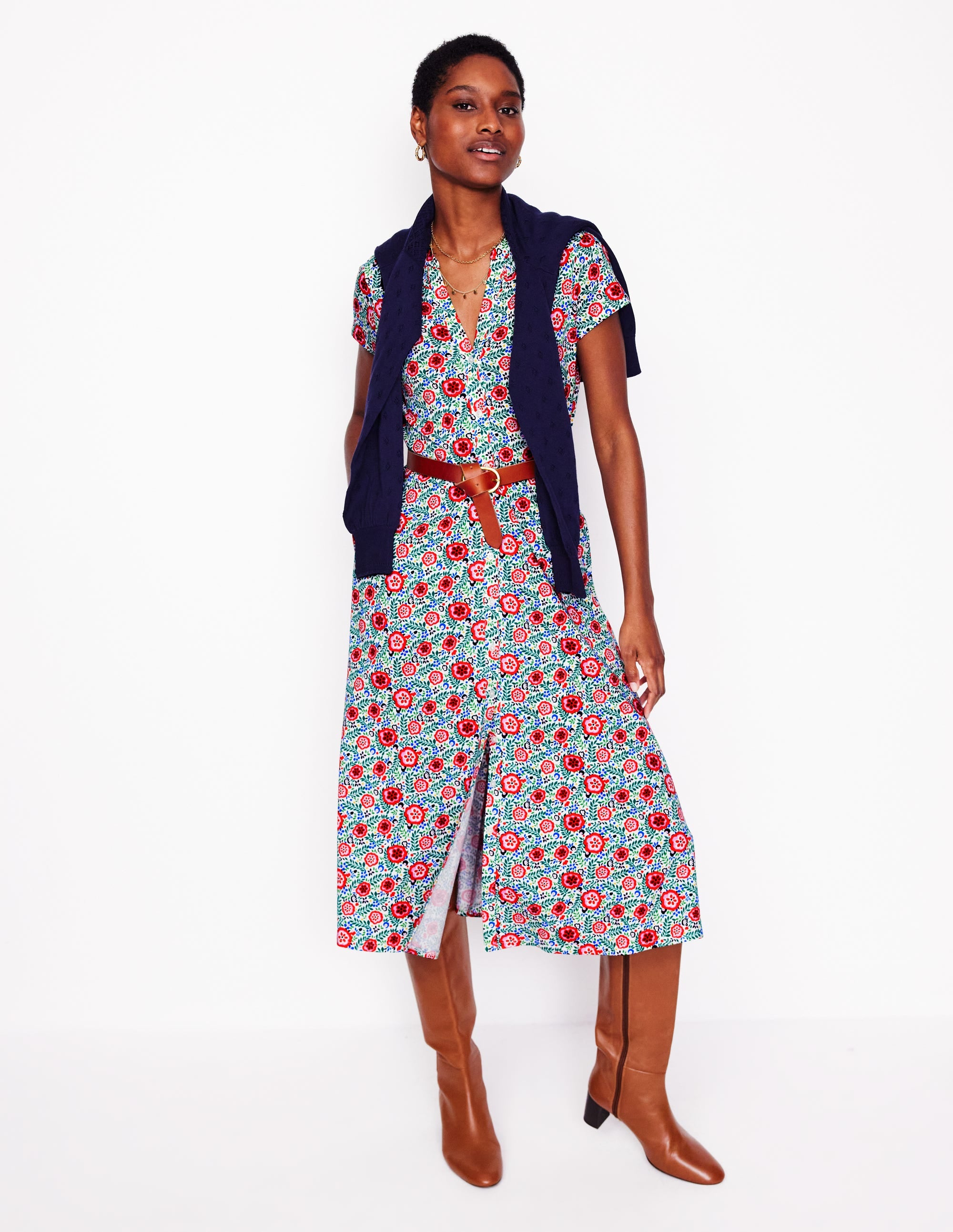 A woman wearing a multicolour foliage scatter print shirt dress with short sleeves, a smocked waist, side pockets, and brown knee-high boots.