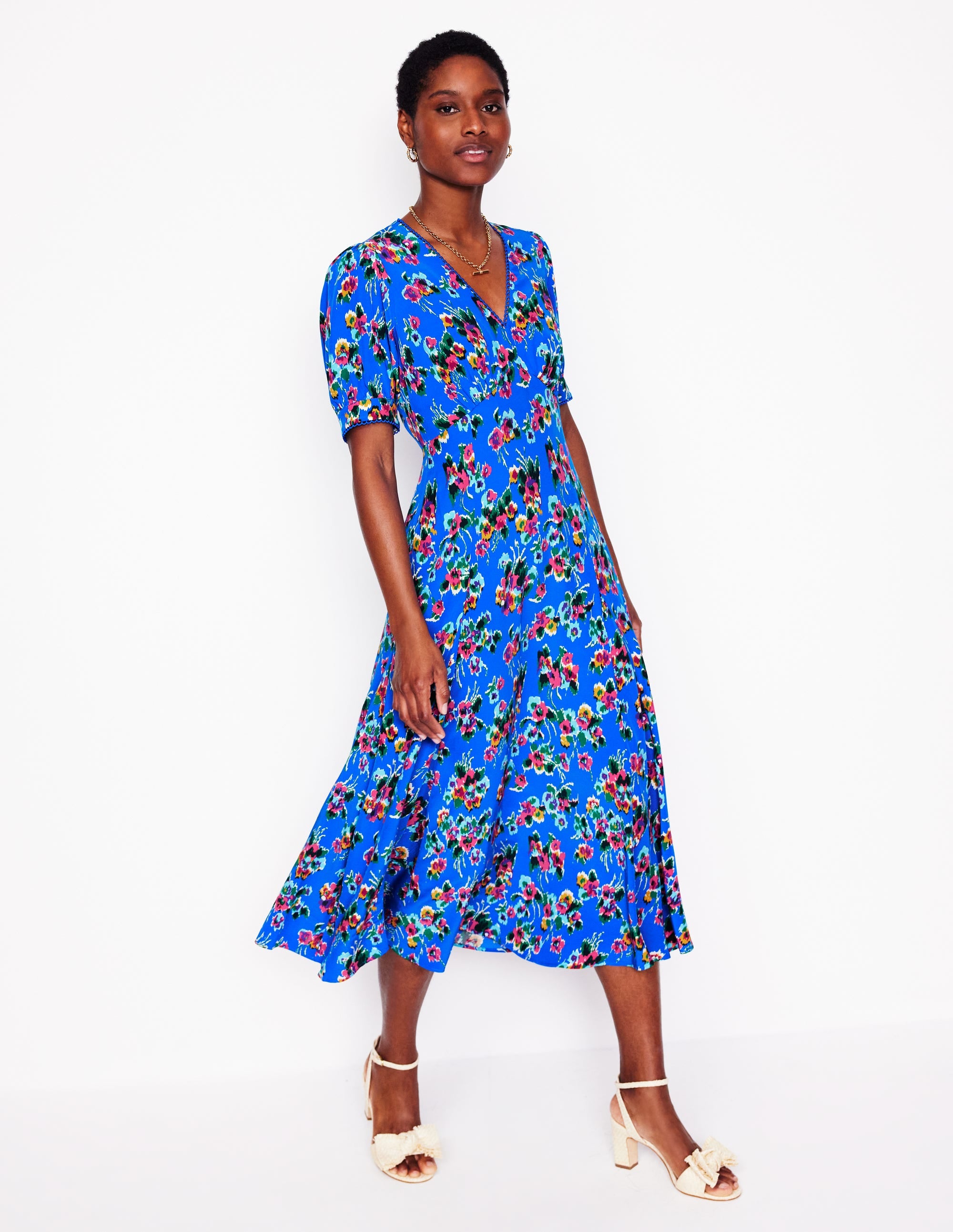 A woman wearing a blue floral tea dress with short puffed sleeves, V-neckline, and mid-calf length, standing against a white background.