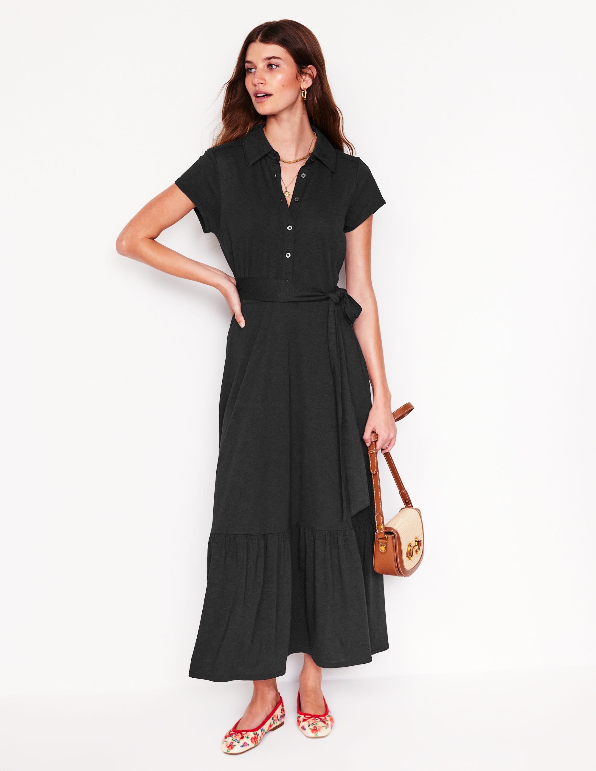A woman wearing a black shirt dress with a collar, short cap sleeves, button-down front, a tie at the waist, and a tiered skirt that reaches below the knees.