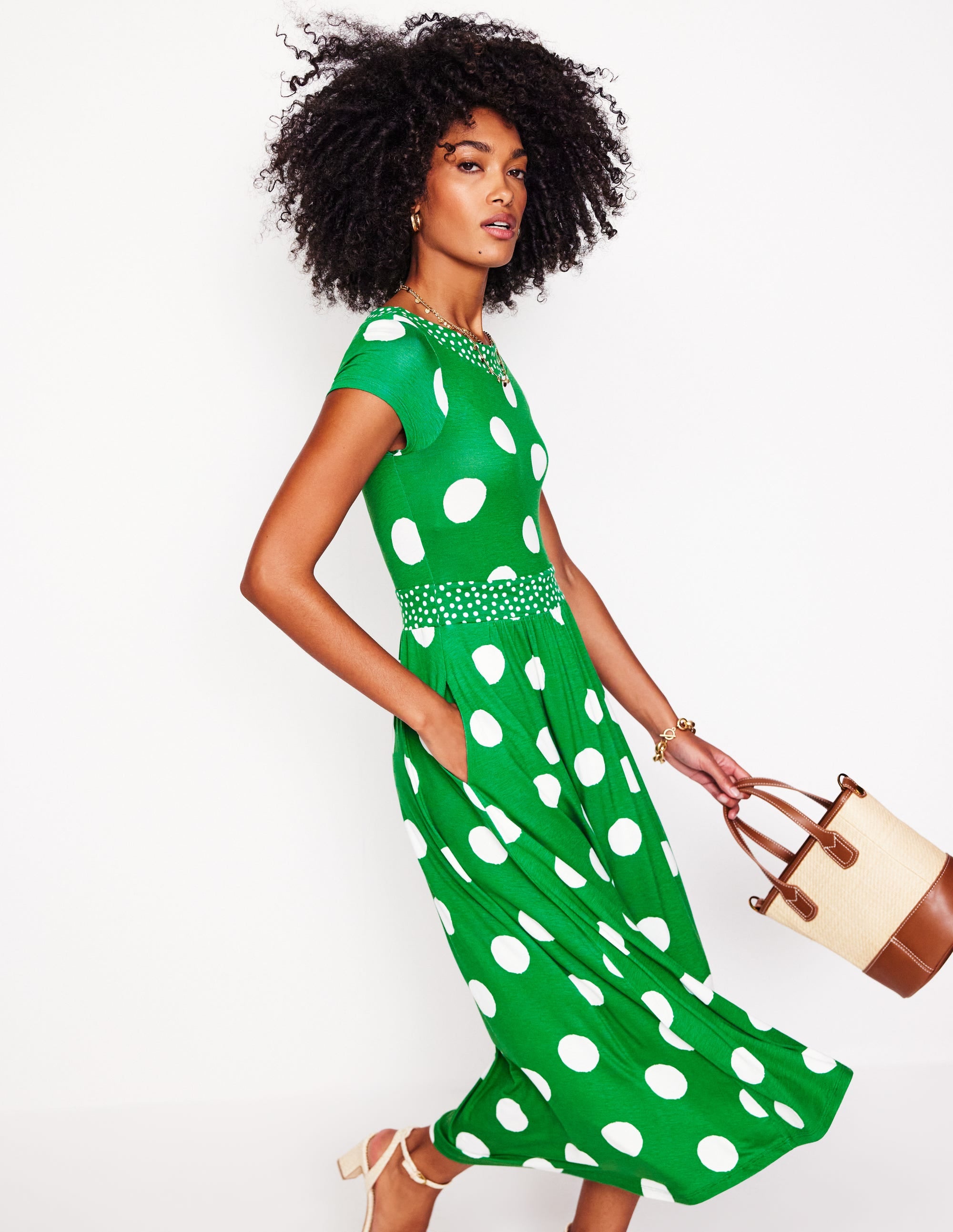 A woman wearing a green midi dress with white abstract dots, short sleeves, and a flared skirt, holding a beige and brown handbag.
