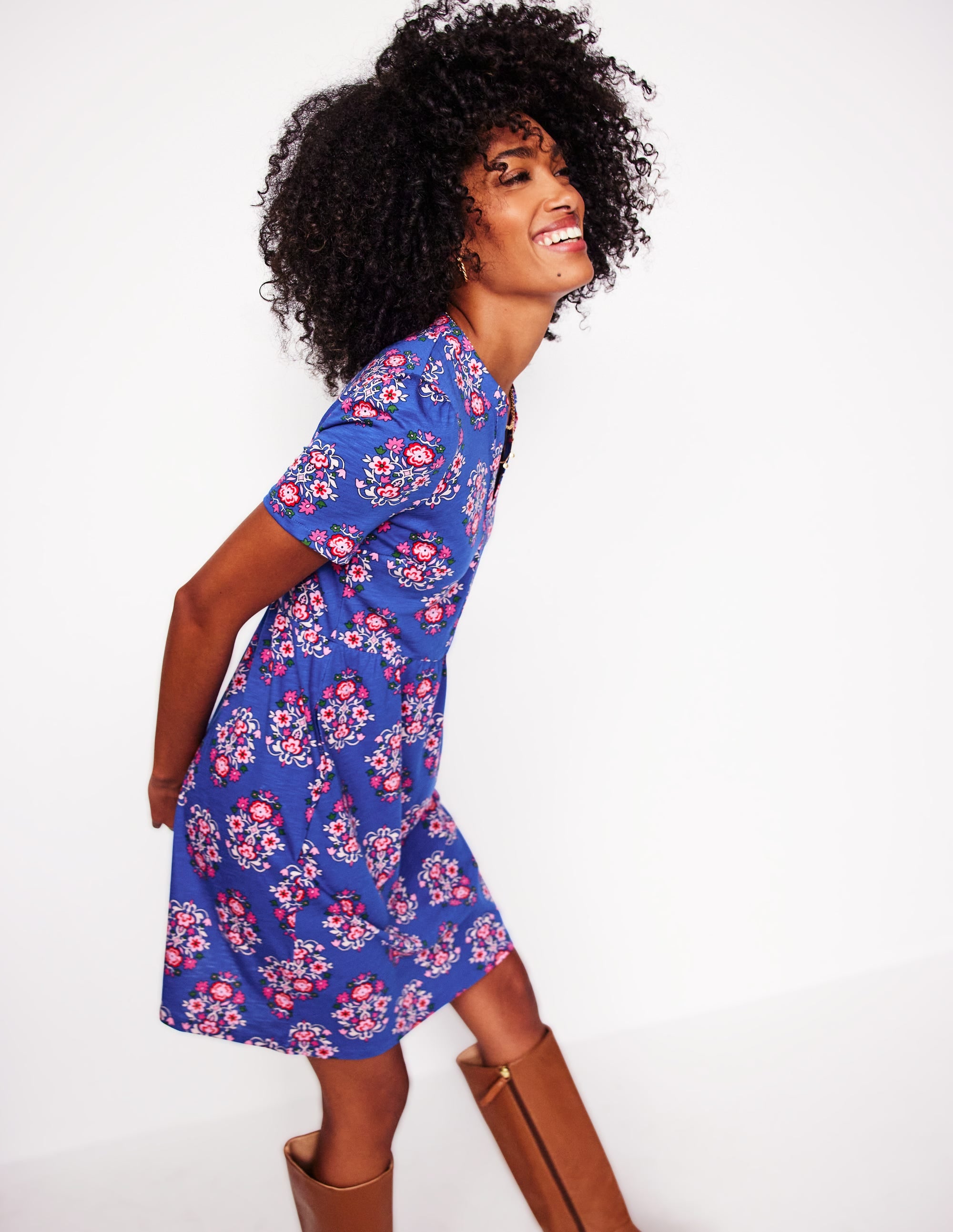 A woman wearing a bright blue short sleeve dress with a wildflower stamp pattern, featuring a gathered waist and a fit and flare silhouette, standing against a white background.