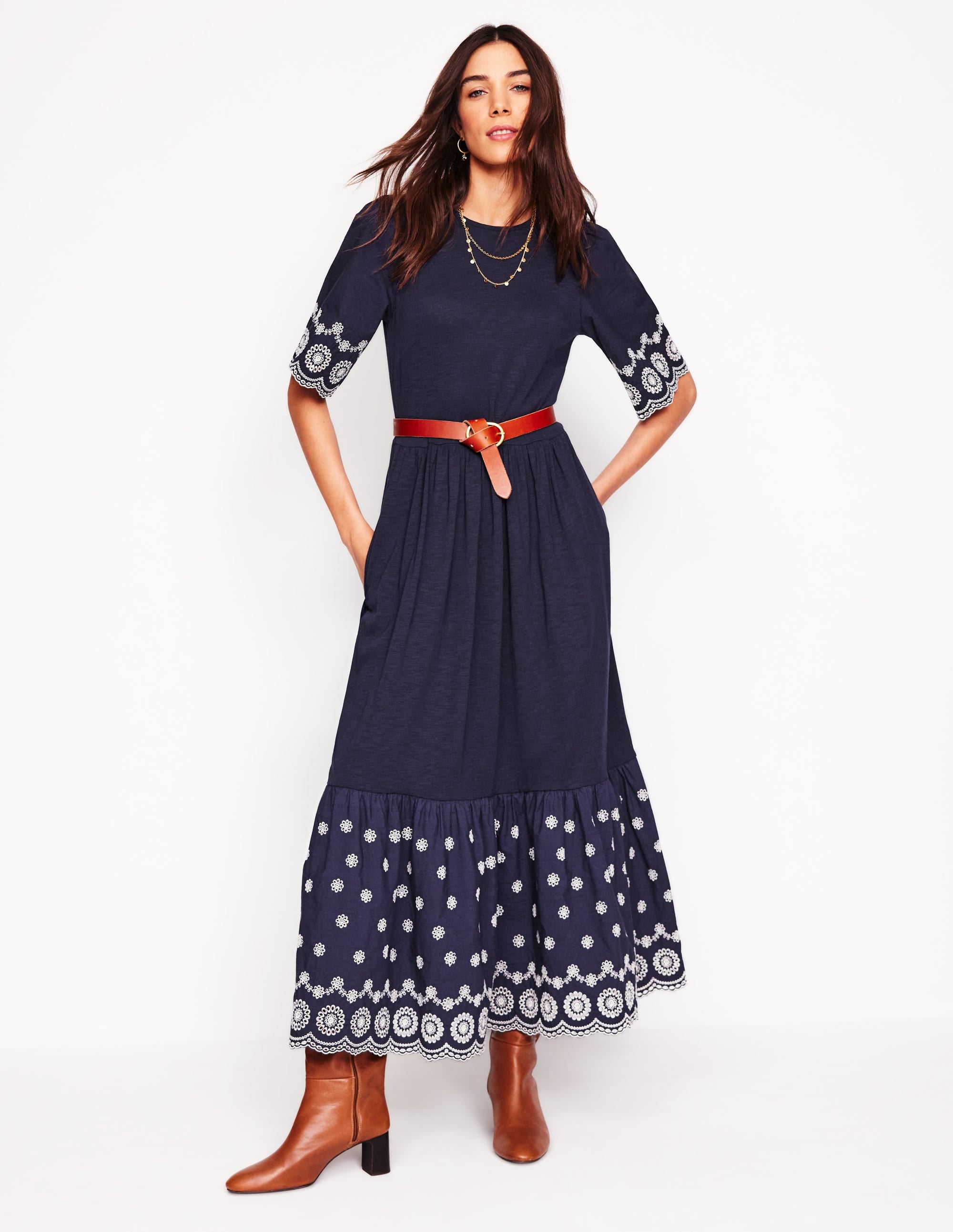 A woman wearing a navy broderie embroidered maxi dress with a fit and flare silhouette, side pockets, and a ruffled hem with white embroidery details.