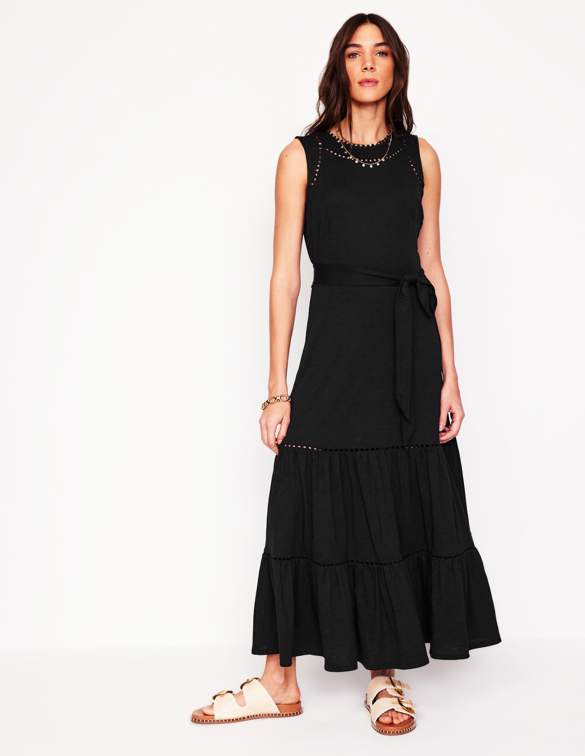 A woman wearing a sleeveless black tiered maxi dress with a waist belt, pom-pom trim accents, and side pockets.
