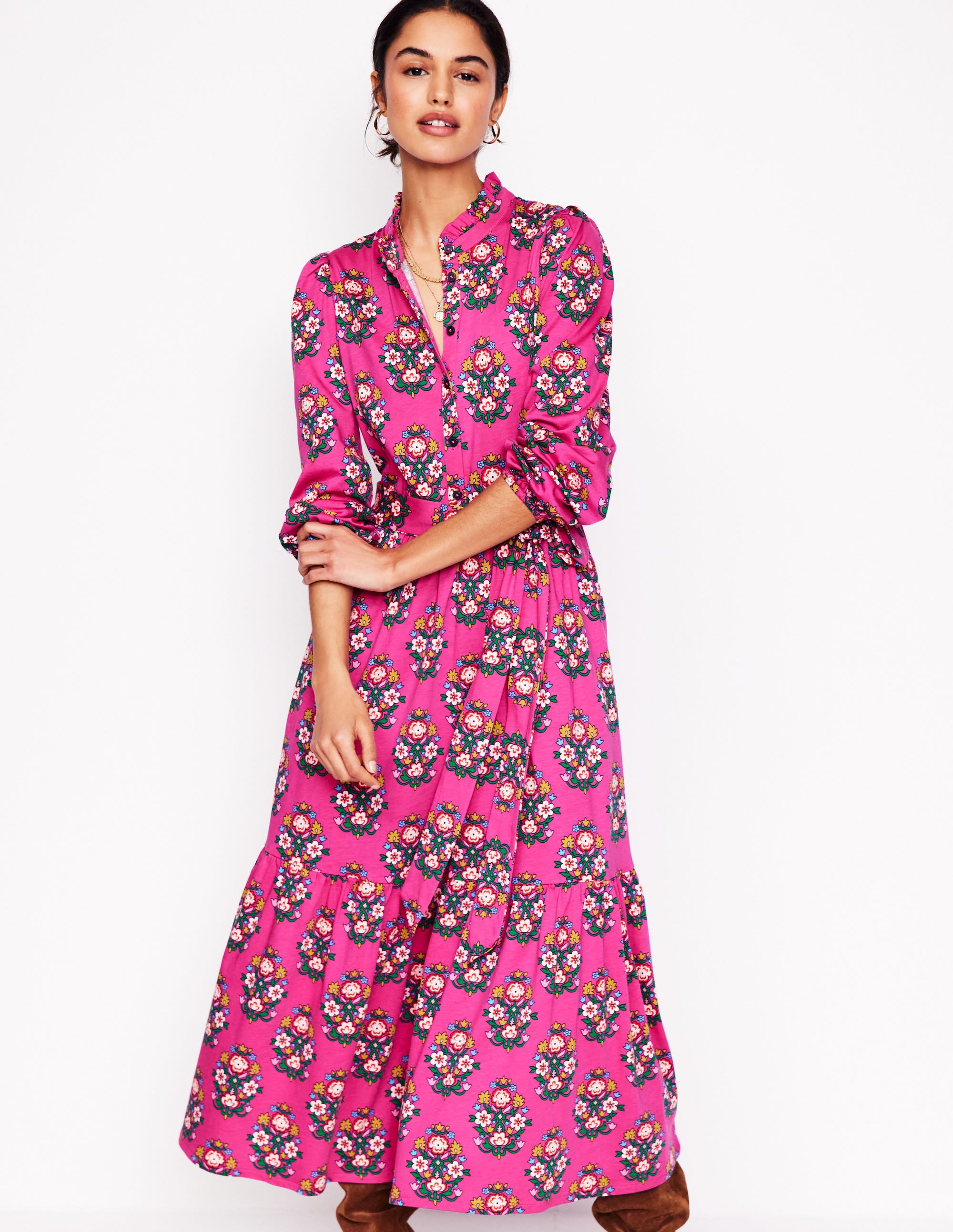 A woman wearing a pink floral tiered maxi dress with long sleeves, a collar, and a belt, standing against a white background.
