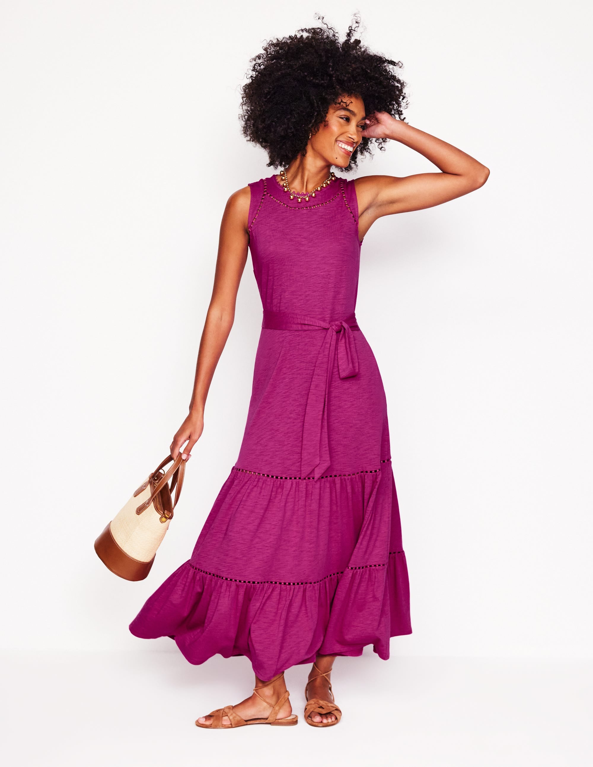 A woman wearing a sleeveless, fuchsia tiered maxi dress with pom-pom trim, a waist belt, and side pockets, standing against a white background.