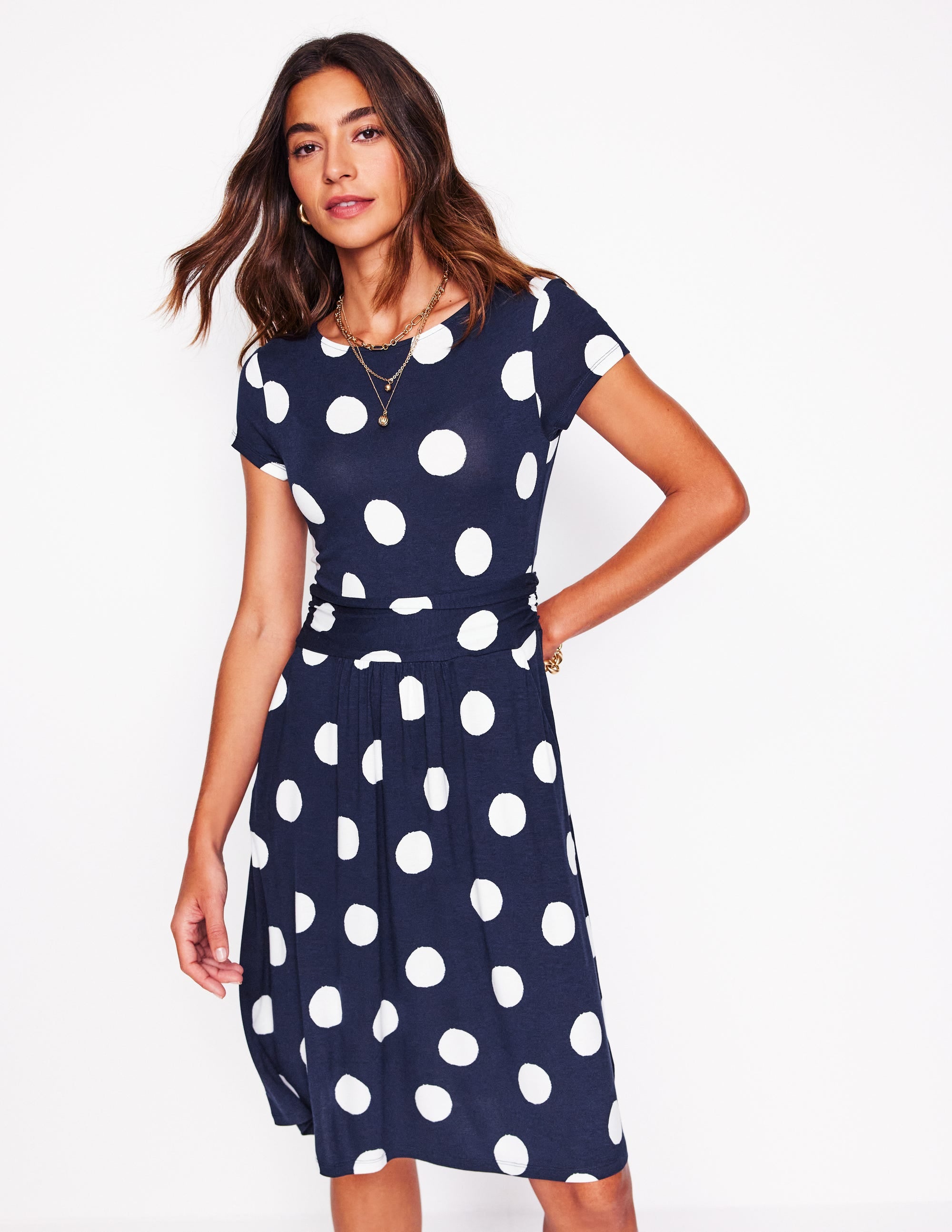 A woman wearing a navy blue dress with large white abstract dots, short sleeves, and a fitted waist, standing against a white background.