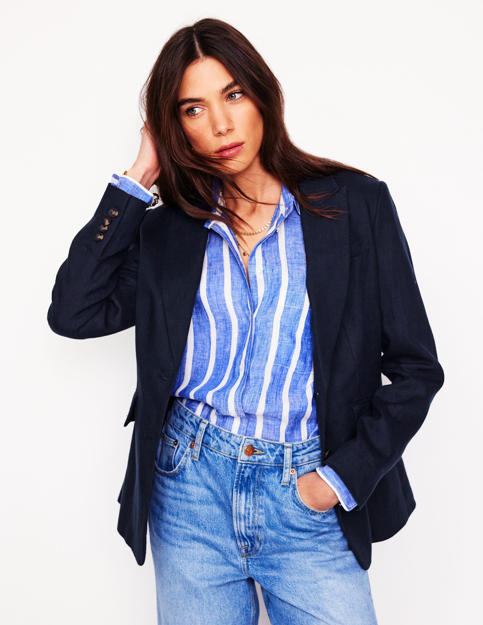 A woman wearing a navy linen blazer over a blue and white striped shirt, with her hand touching her hair.
