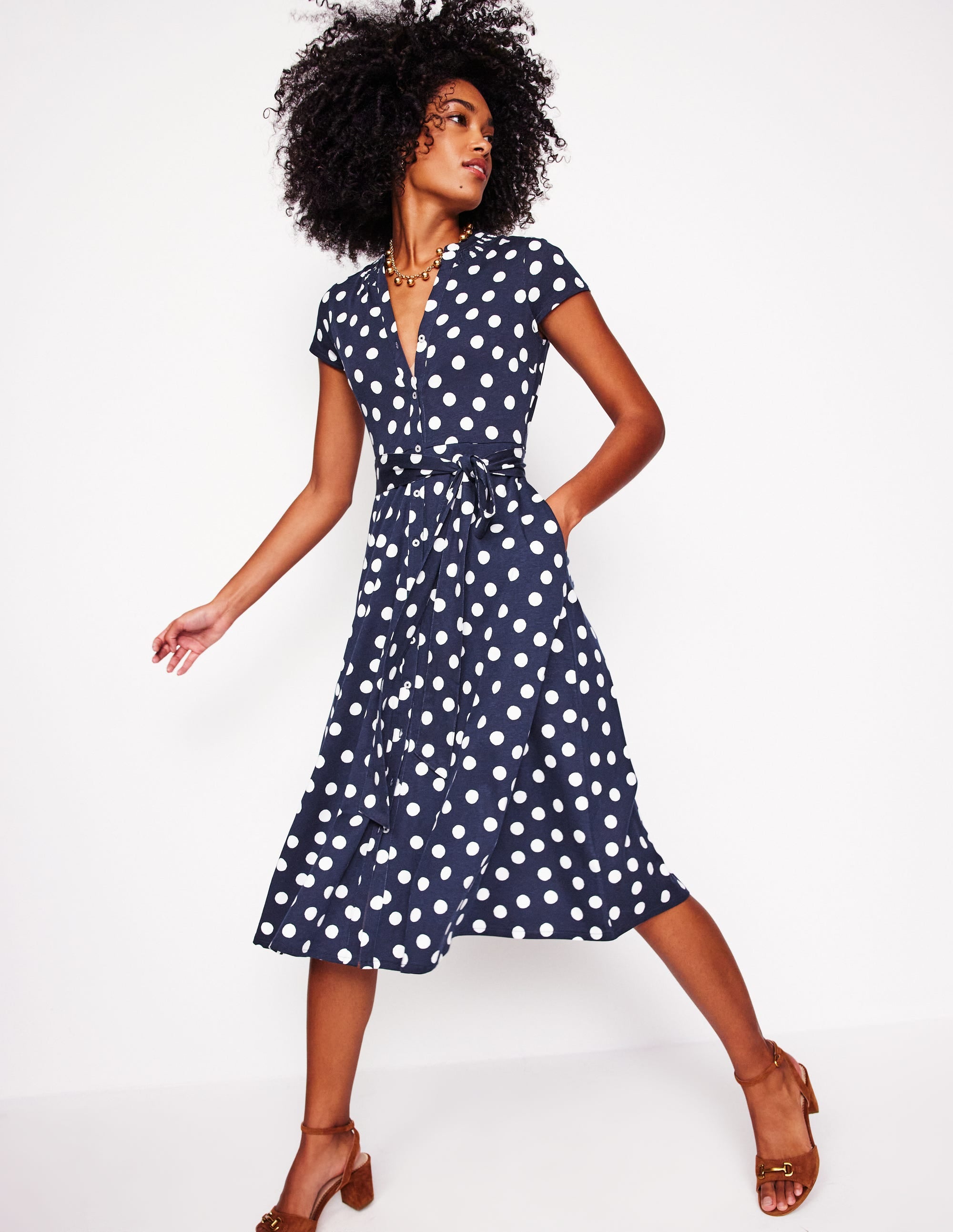 A woman wearing a navy blue shirt dress with white abstract dots, short sleeves, a tie belt at the waist, and side pockets.