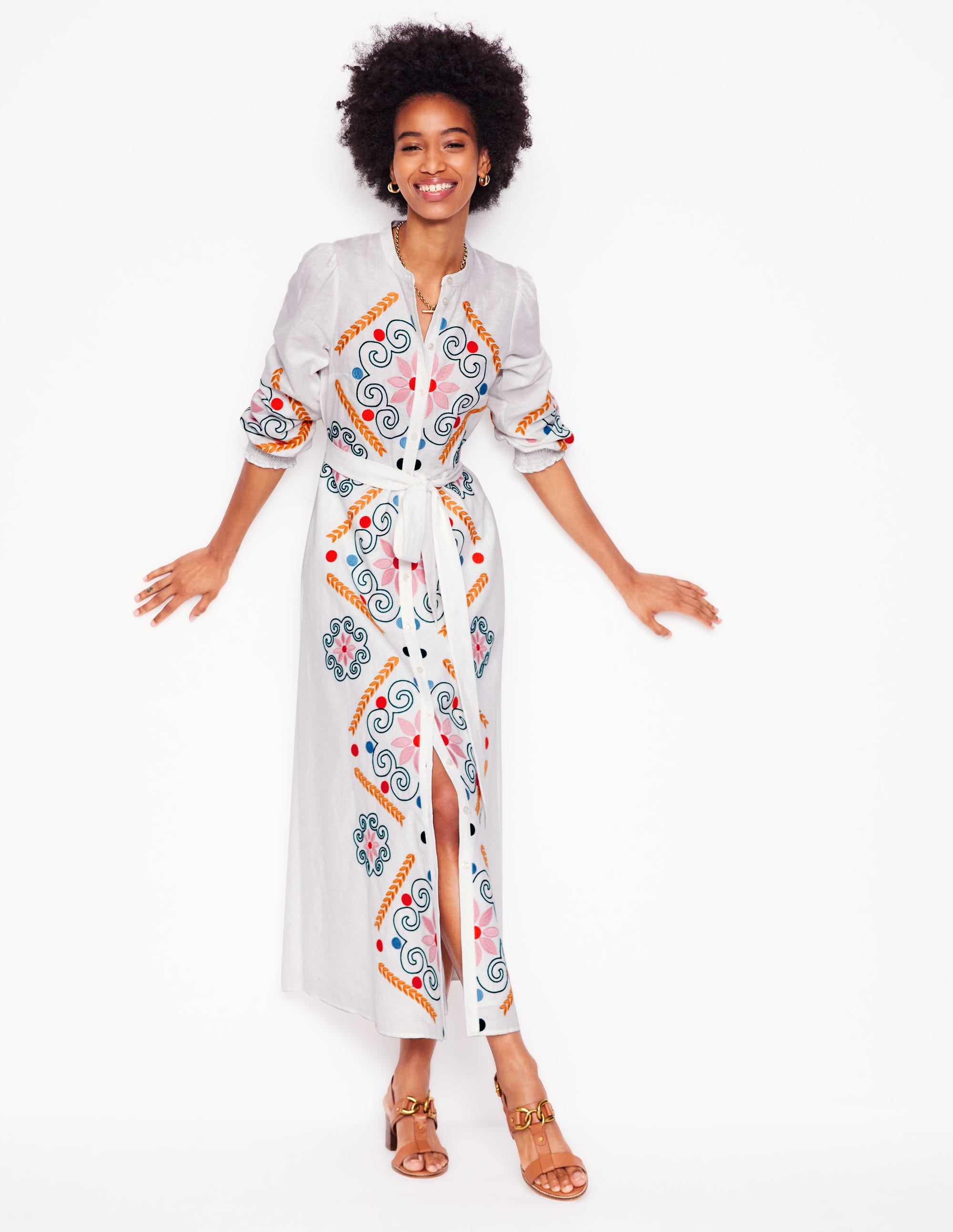 A woman wearing a white linen blend shirt dress with colorful embroidery, full skirt, pockets, smocked cuffs, and a tie waist belt.