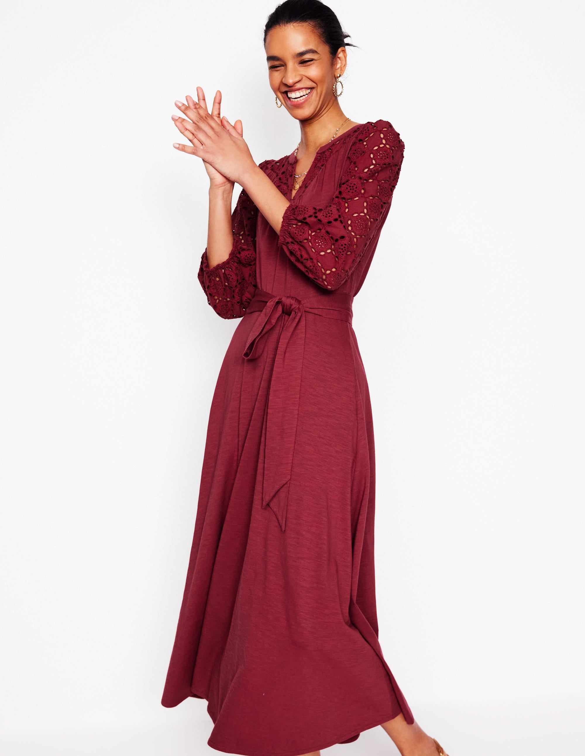 A woman wearing a garnet red, long, relaxed-fit dress with eyelet embroidery on the sleeves and a waist tie, smiling and standing against a white background.