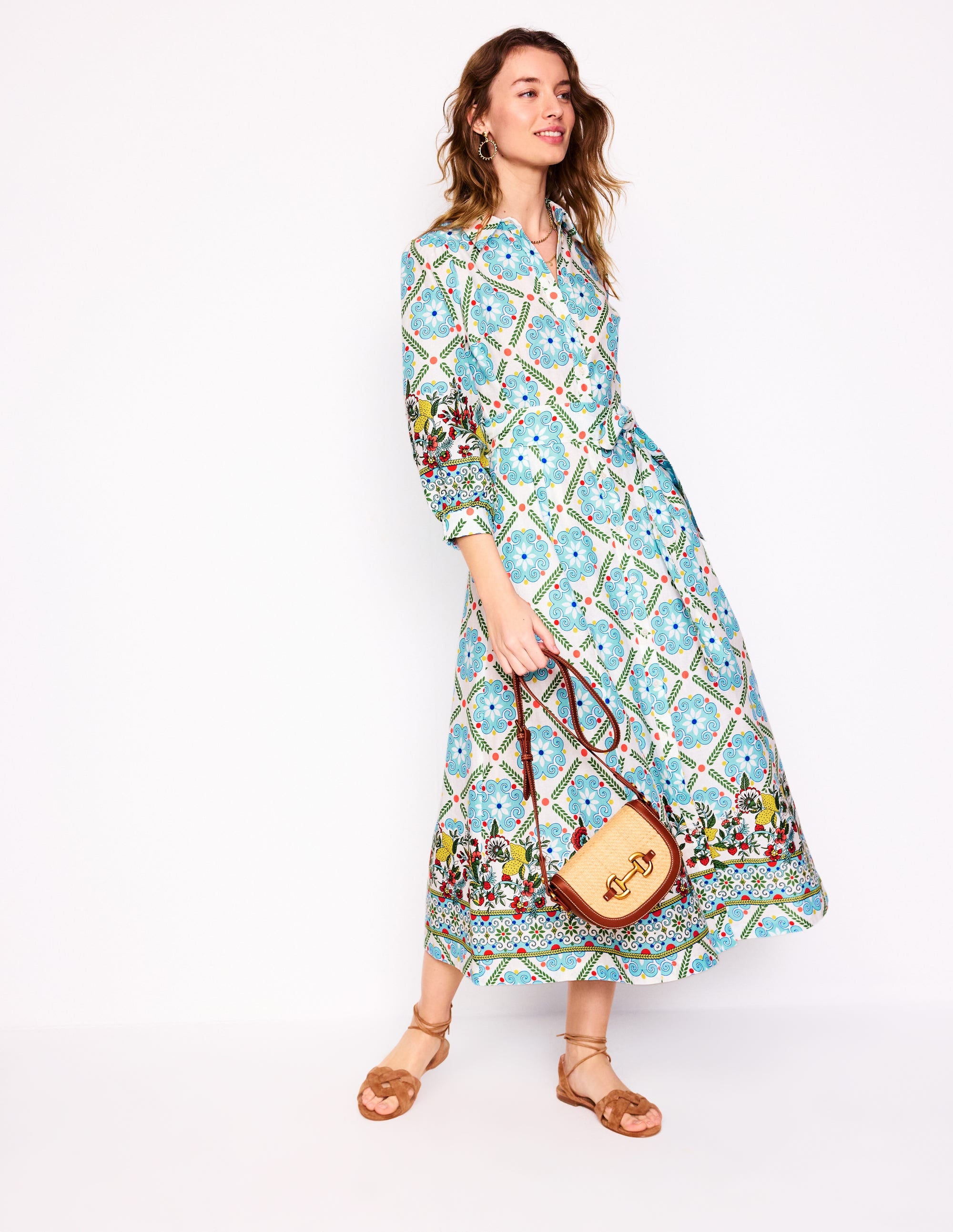 A woman wearing a long, colorful patterned shirt dress with a belt, pockets, and a collar, holding a small handbag.