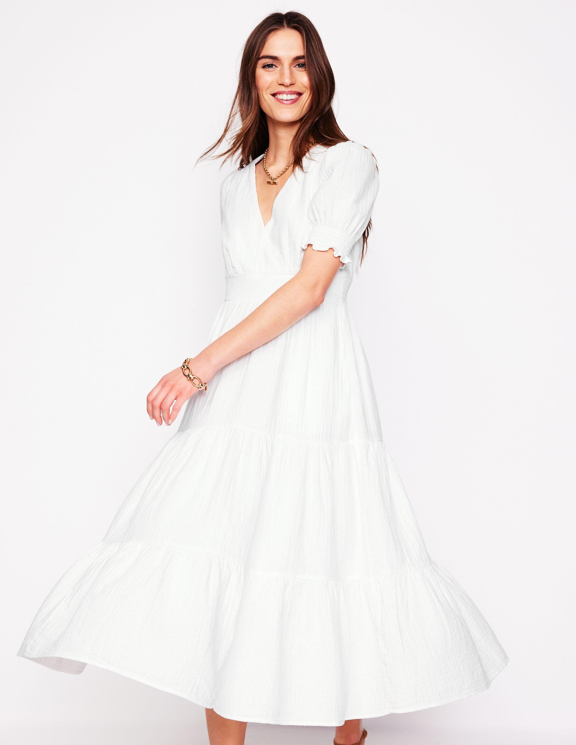 A woman wearing a white midi dress with a tiered skirt, short puffed sleeves, and a V-neckline, standing against a plain background.