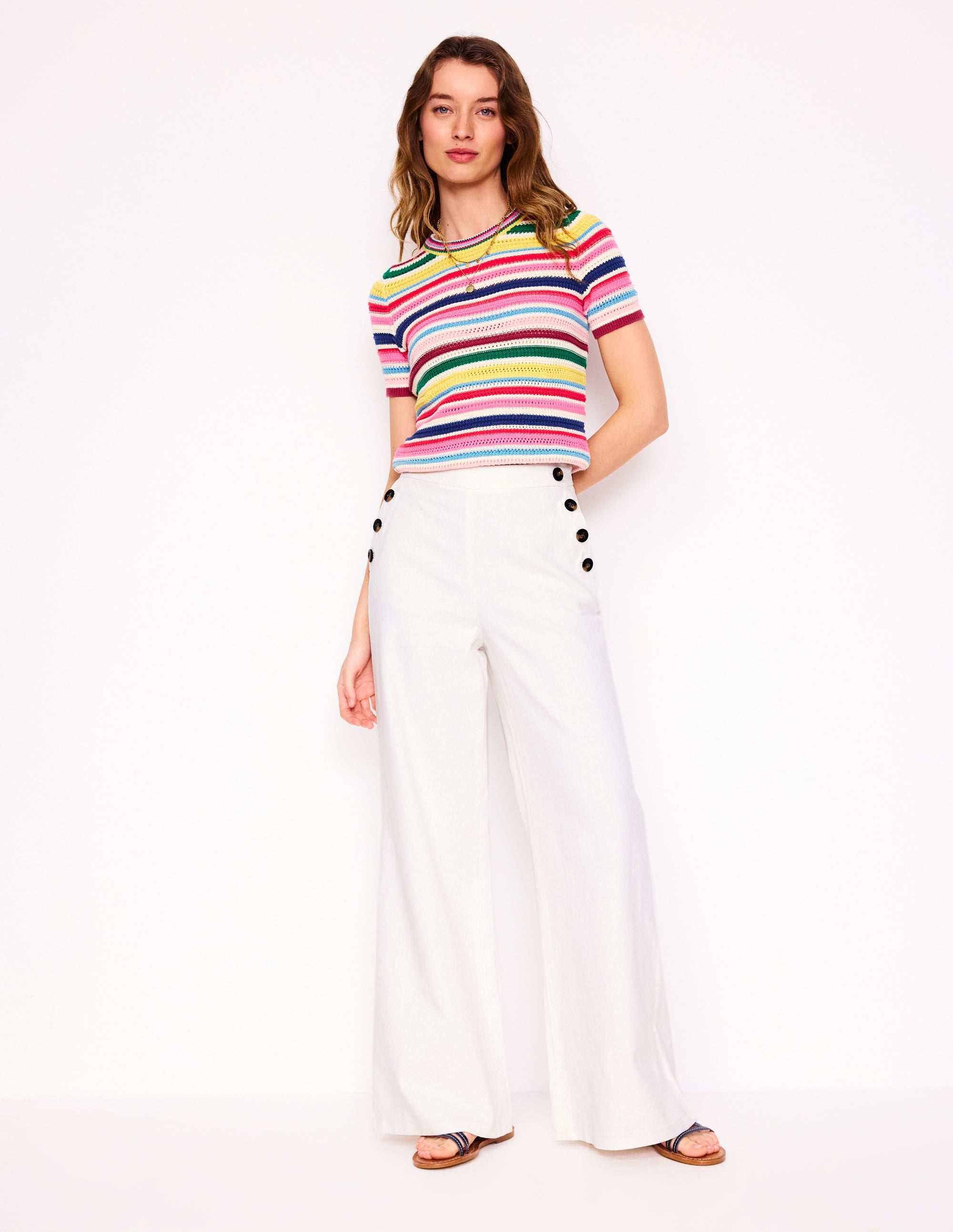 A woman wearing white high-waisted sailor trousers with wide legs, buttoned bib front, and side zip, paired with a colorful striped top.