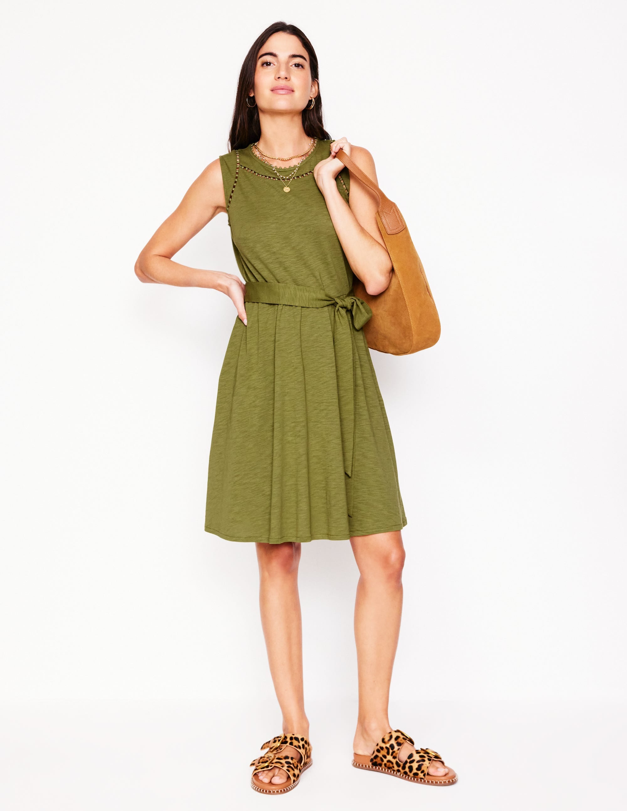A woman wearing a sleeveless deep olive green jersey dress with a tiered skirt, pom-pom trim, and a detachable belt, standing against a plain background.