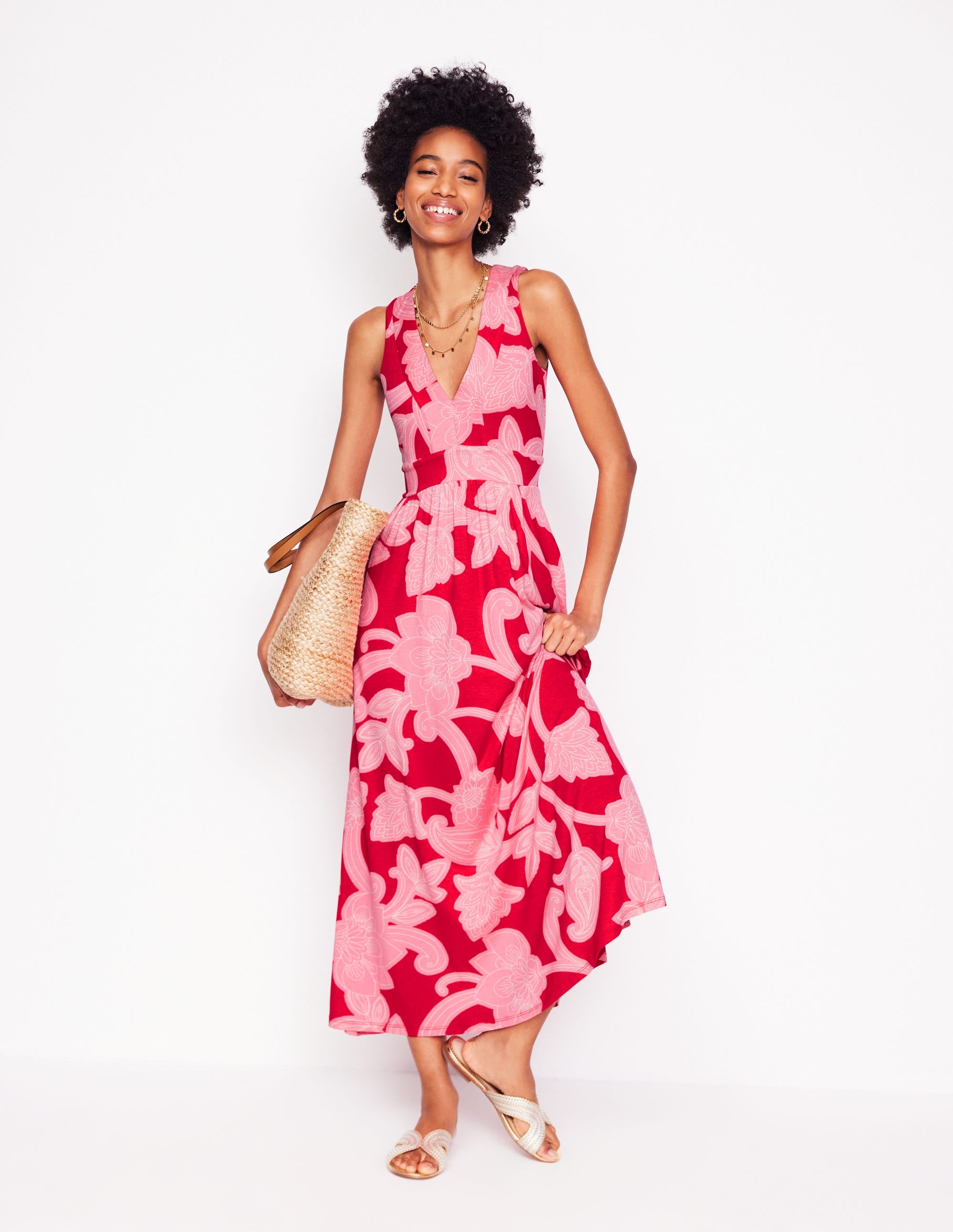 A woman wearing a sleeveless, V-neck maxi dress with a red and pink paisley whirl print, tiered design, and side pockets, standing against a white background.