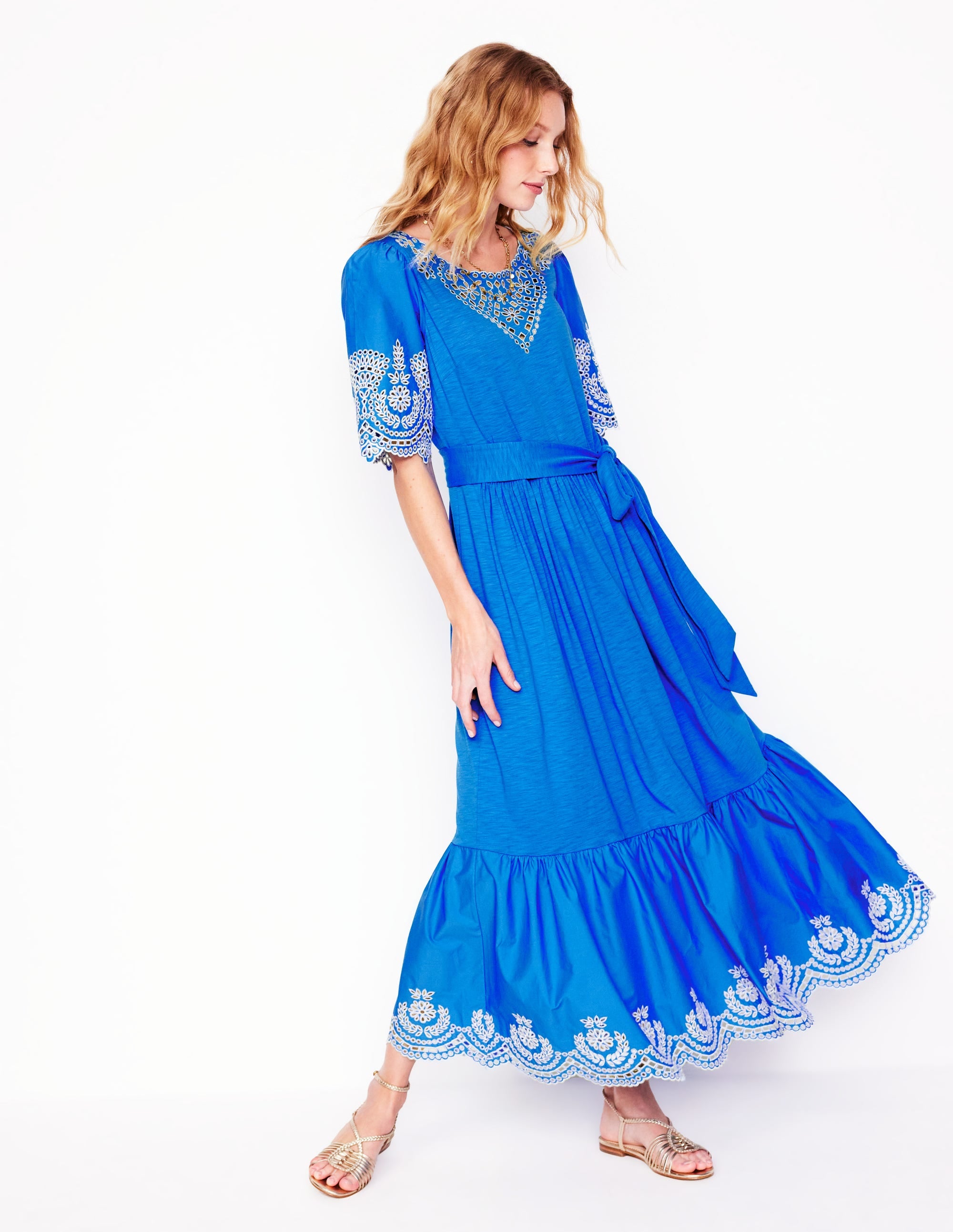A woman wearing a bright cerulean blue maxi dress with embroidered details on the sleeves and hem, featuring a fit and flare silhouette, side pockets, and a waist tie.
