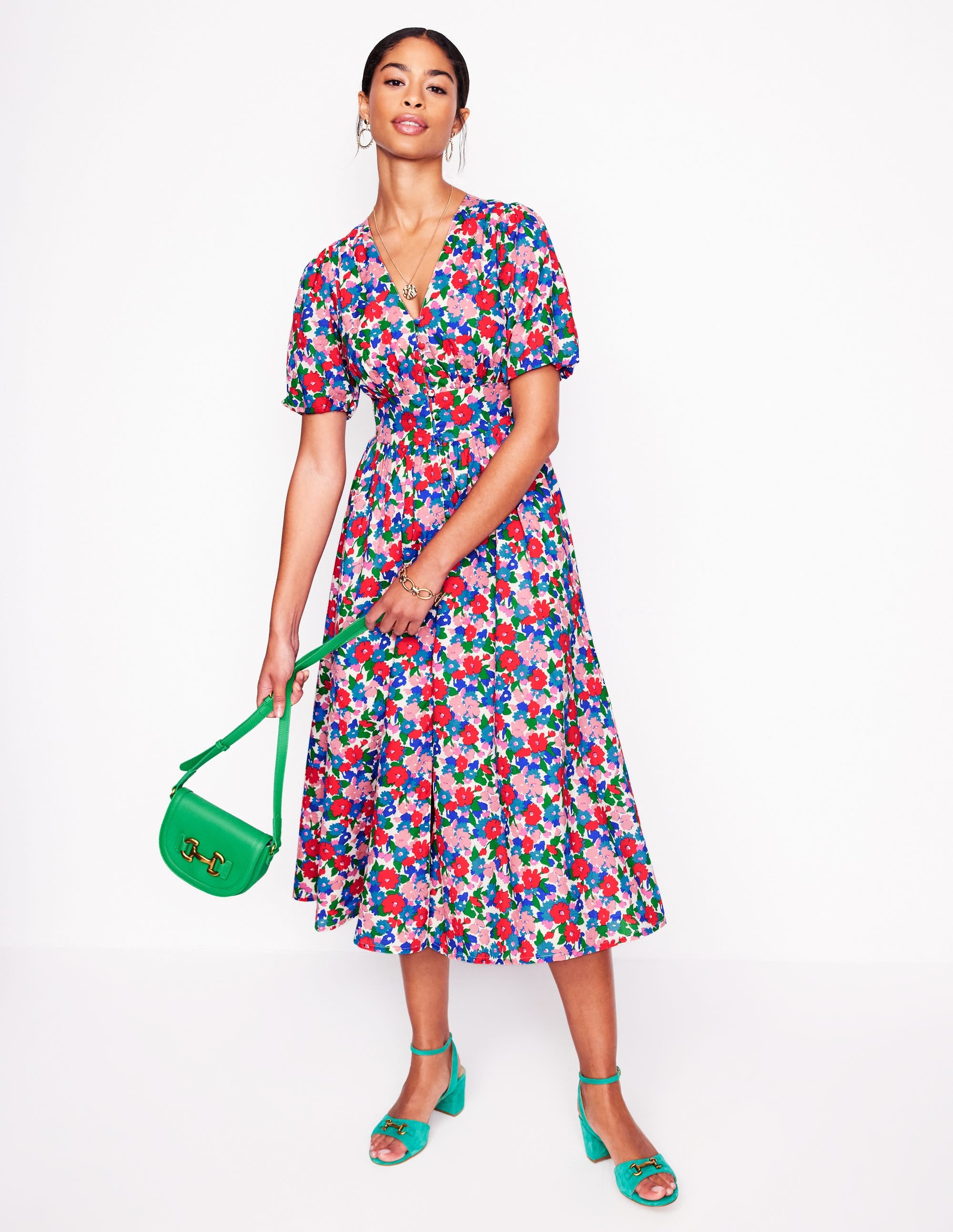 A woman wearing a colorful floral midi dress with puff sleeves and a fitted waist, holding a green handbag, standing against a plain white background.