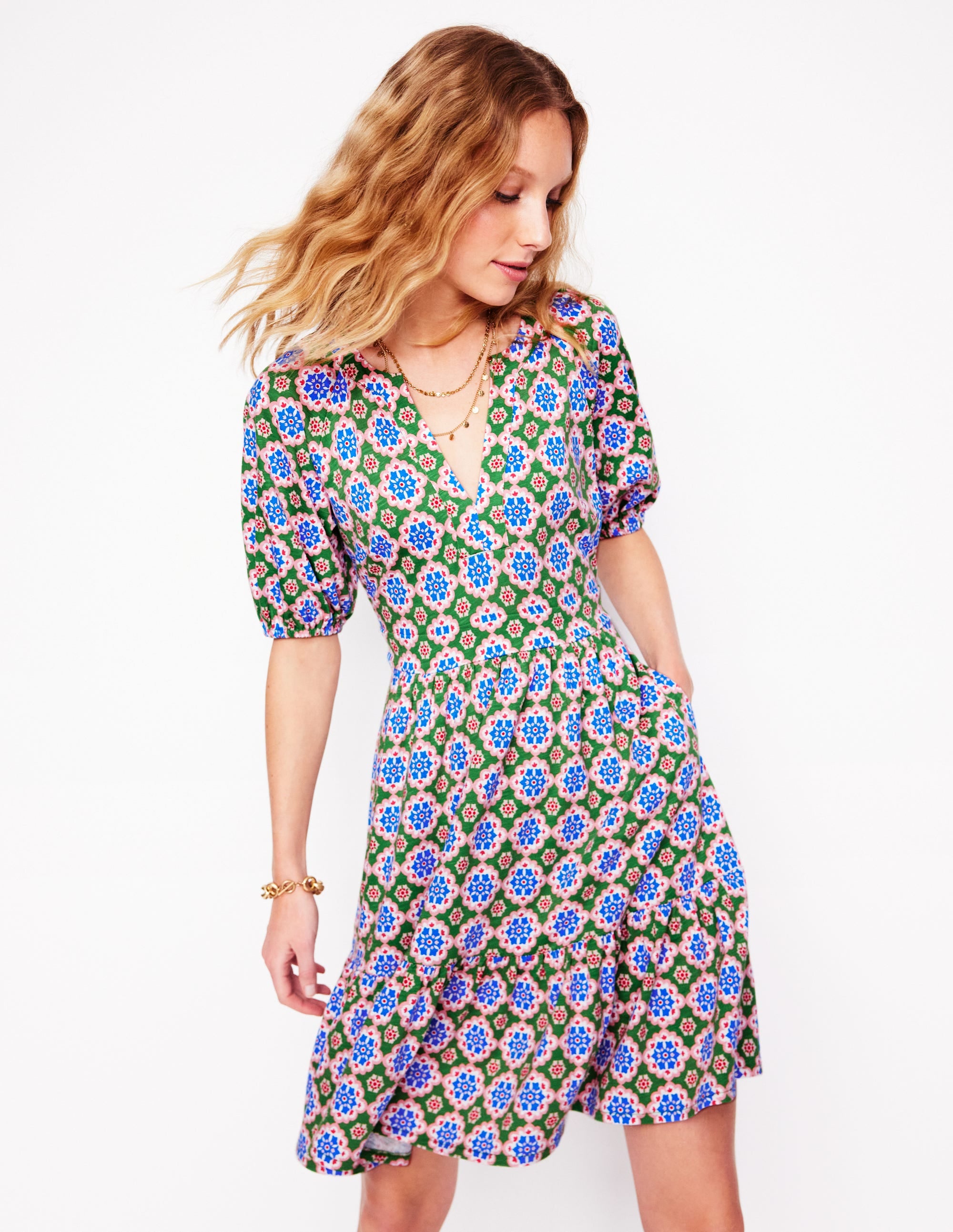 A woman wearing a green floral jersey dress with short puffed sleeves, V-neckline, and side pockets.