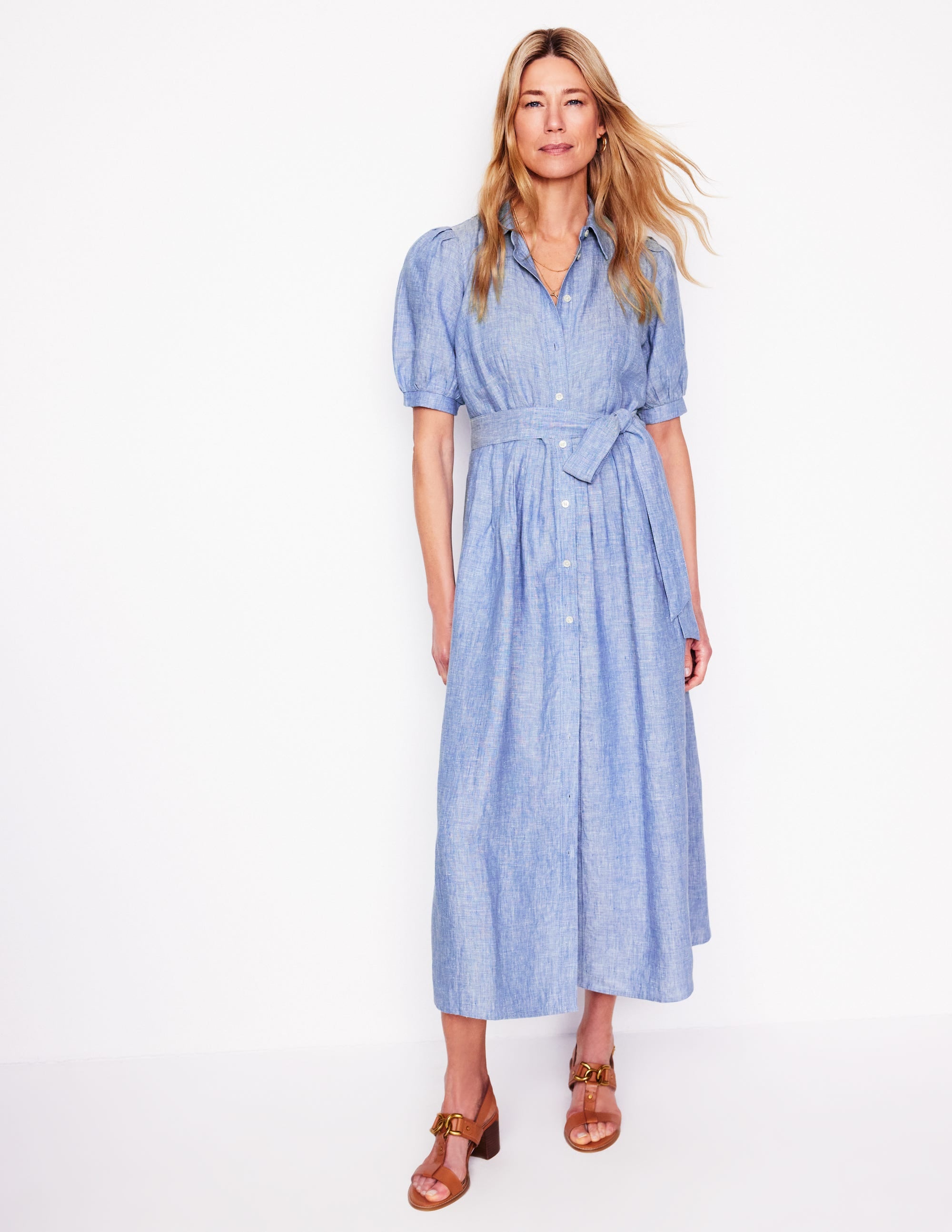 A woman wearing a long, flowing Grey Blue Chambray linen maxi shirt dress with puffed sleeves, a waist tie, and side pockets.