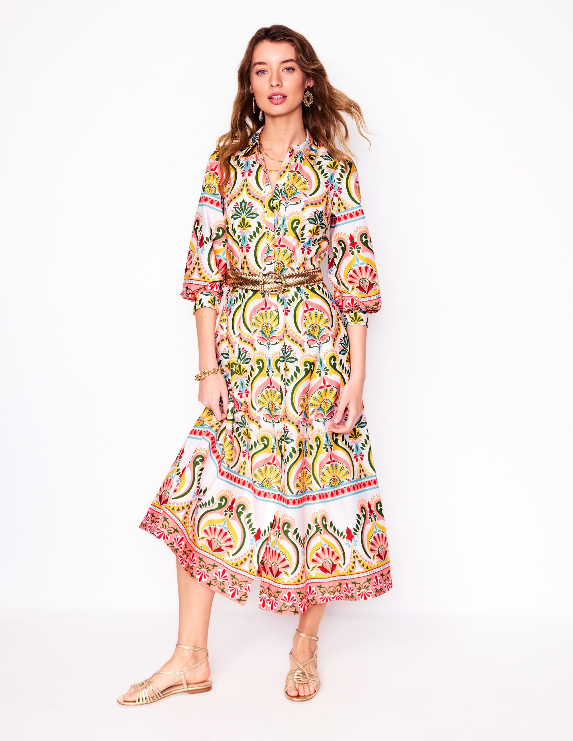 A woman wearing a colorful, floral patterned maxi dress with long sleeves, a belt at the waist, and side pockets.