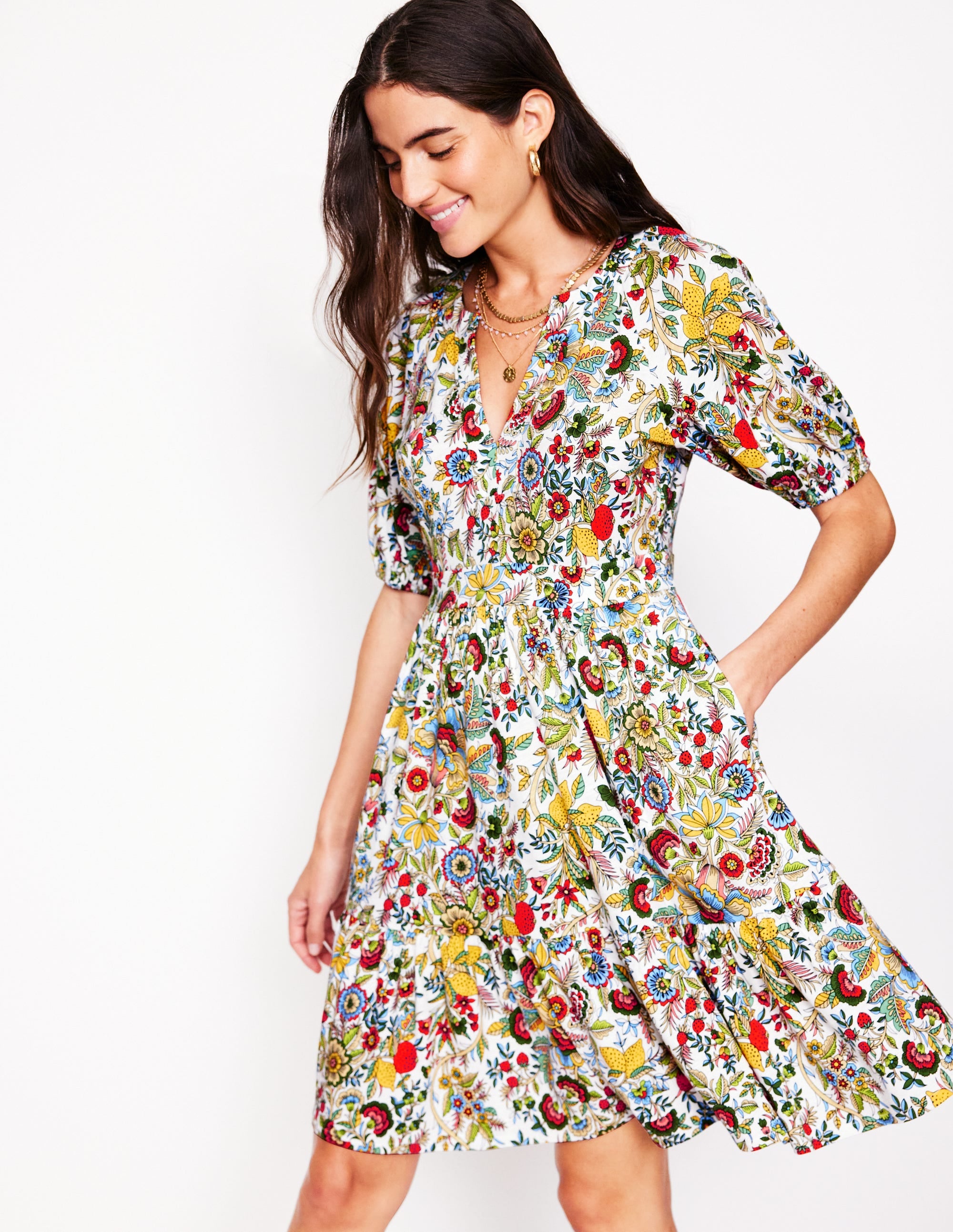 A woman wearing a colorful floral dress with short puffed sleeves, V-neckline, and side pockets, standing against a plain background.