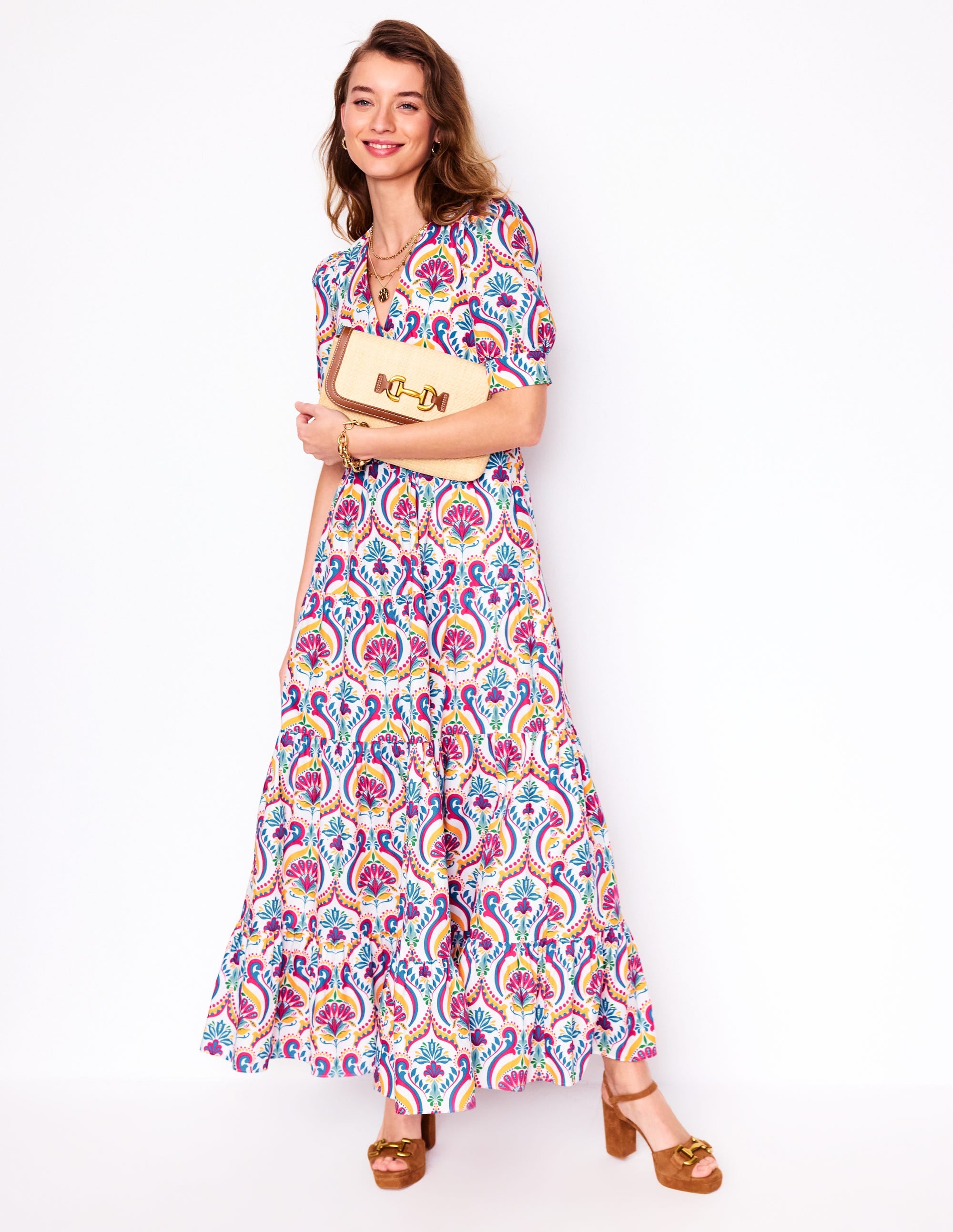 A woman wearing a multicolour floral maxi dress with puff sleeves, tiered skirt, and V-neckline, holding a beige clutch, standing against a white background.