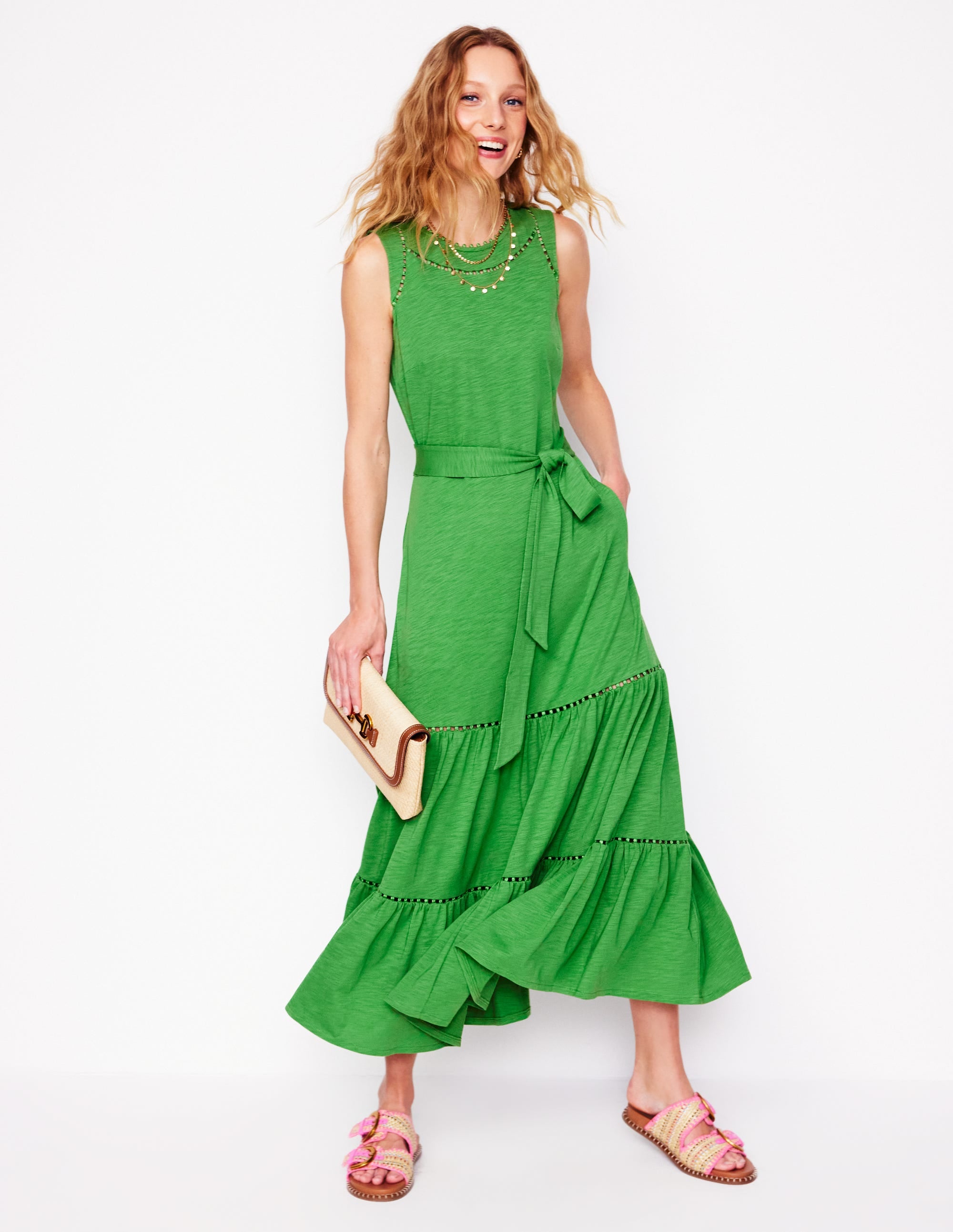 A woman wearing a sleeveless, green tiered maxi dress with pom-pom trim, a waist belt, and side pockets.