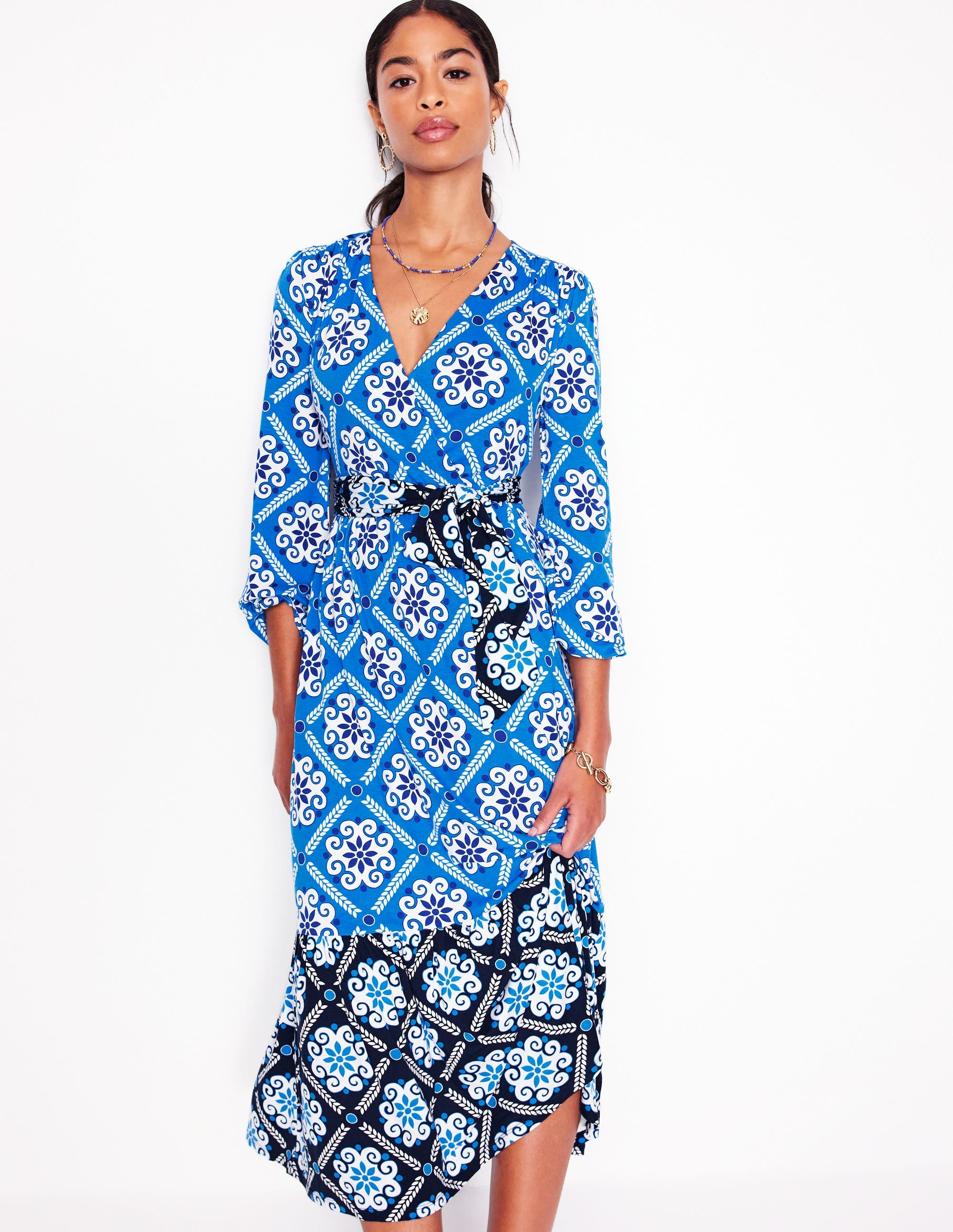 A woman wearing a Cerulean Blue maxi dress with a Baroque Tile pattern, featuring a wrap front, ballooned sleeves, V-neck, and side pockets.