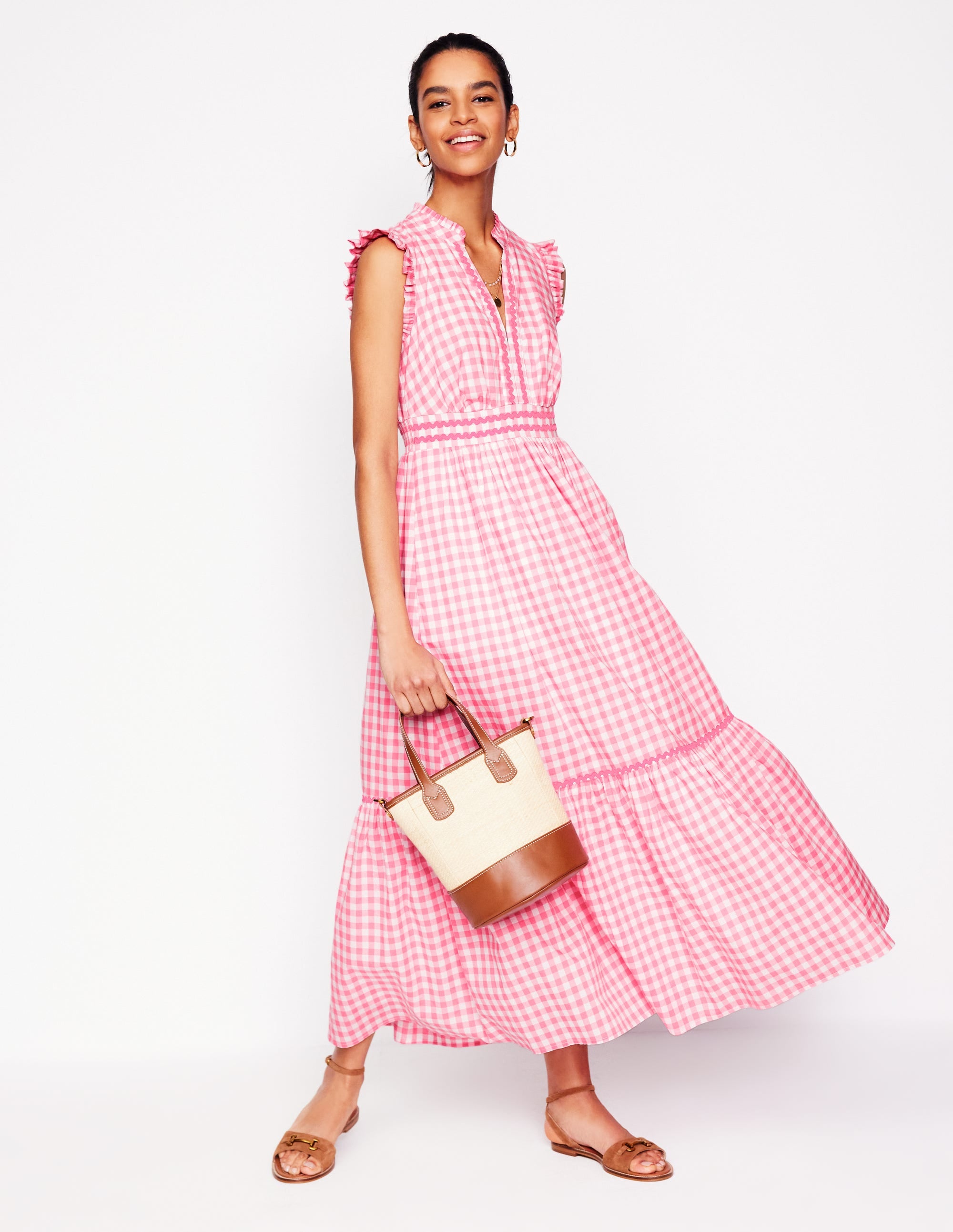 A woman wearing a salmon pink gingham tiered midi dress with ruffled sleeves and a cinched waist, holding a beige and brown handbag.
