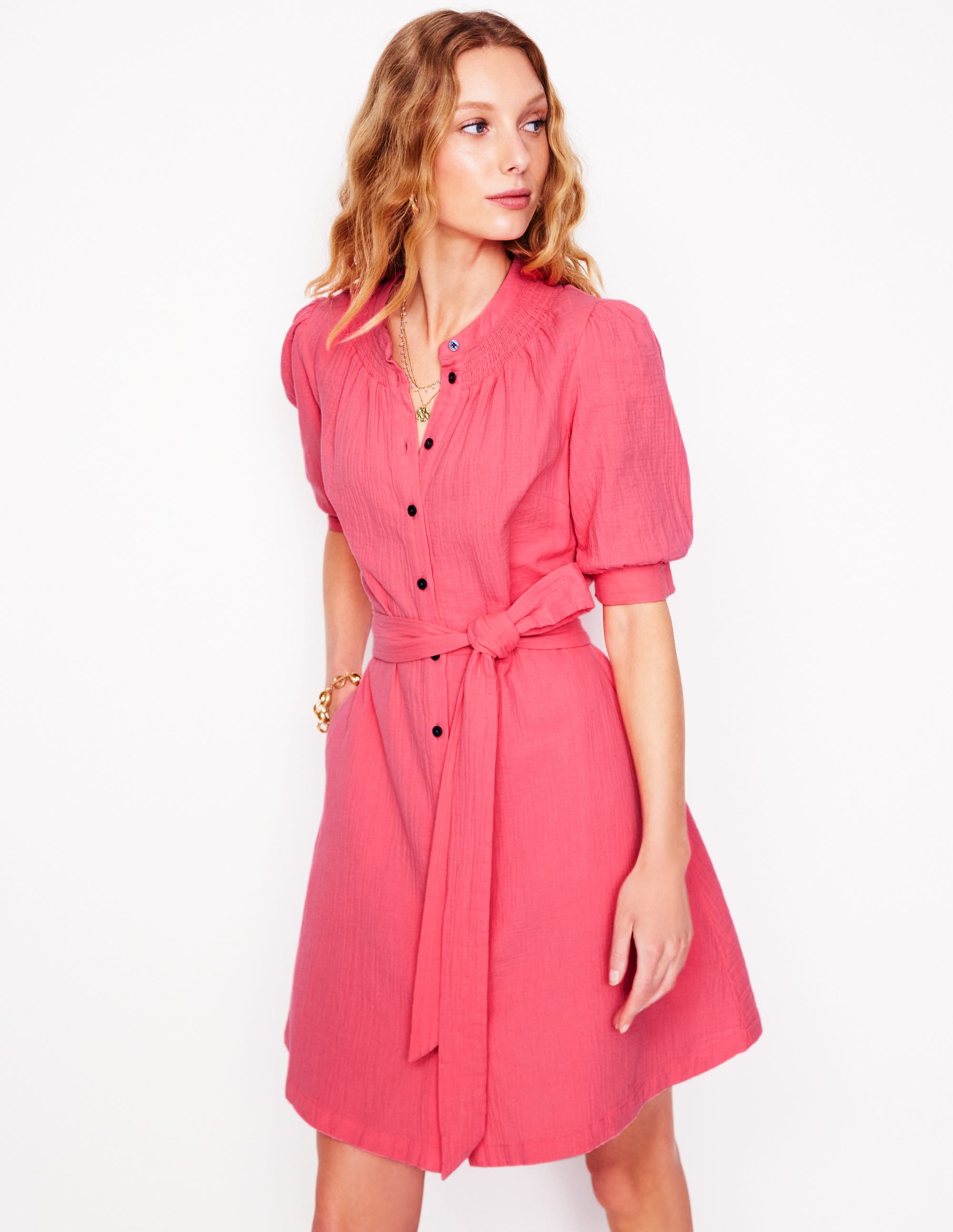 A woman wearing a pink midi dress with gathered sleeves, a smocked neckline, and a belt, standing against a white background.