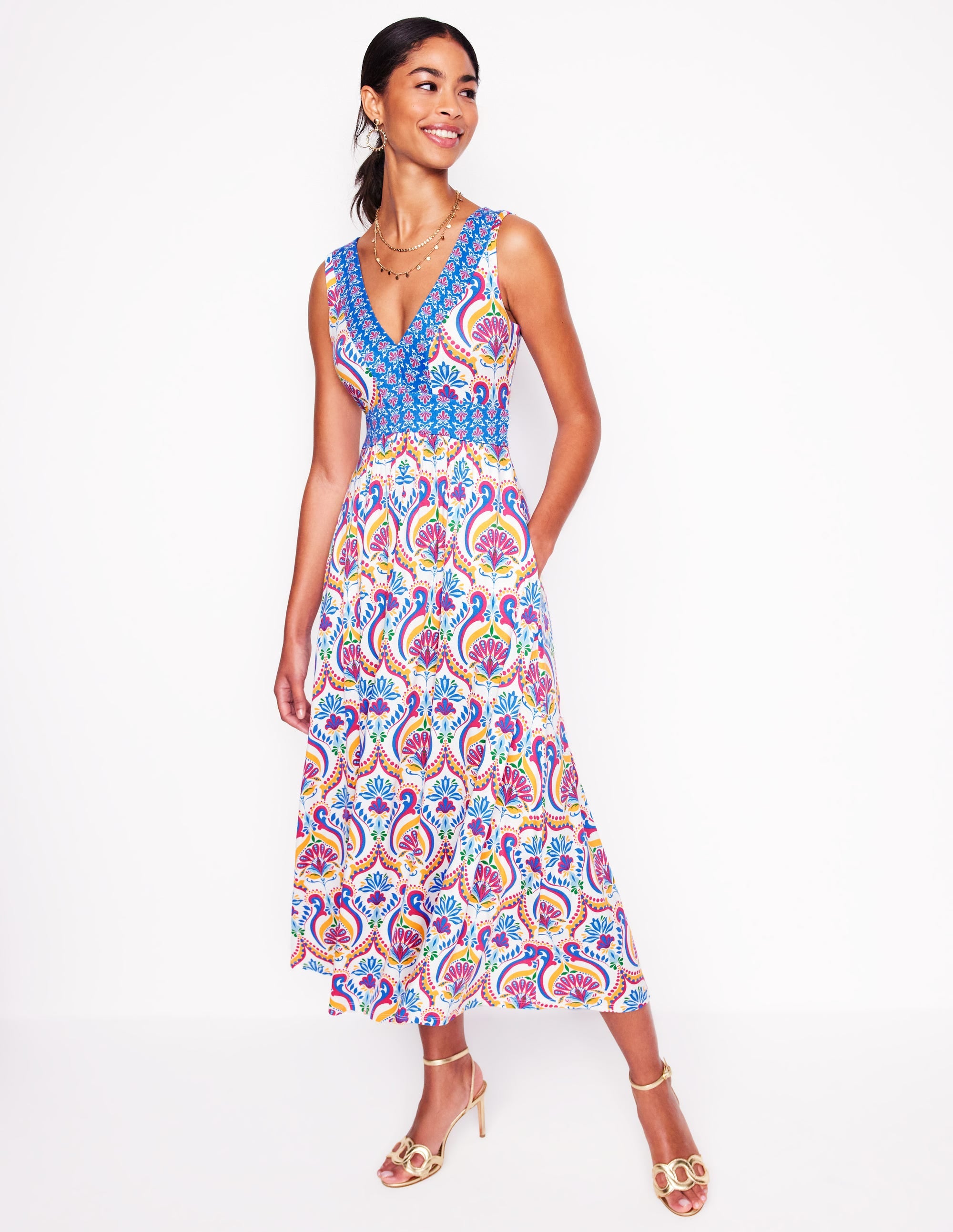 A woman wearing a colorful, sleeveless maxi dress with a tiered design, V-neckline, and floral print, standing against a white background.