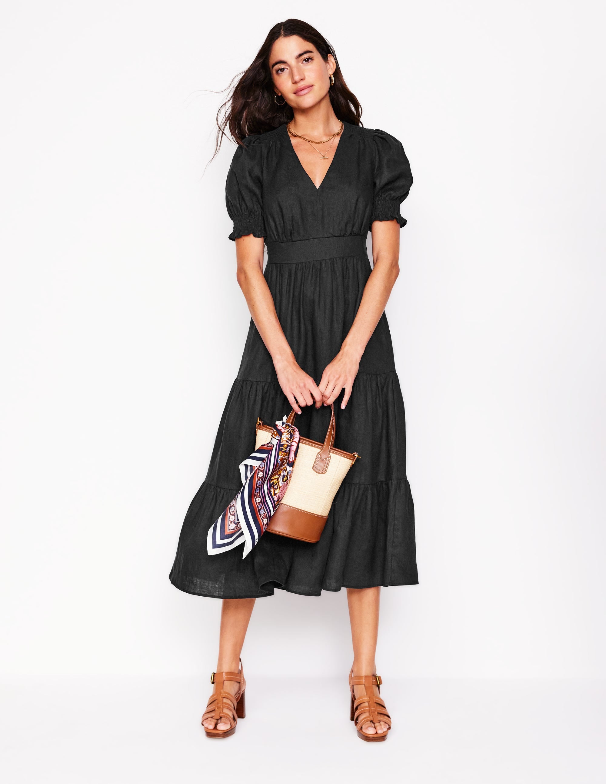 A woman wearing a black linen tiered midi dress with puff sleeves and a V-neck, holding a beige and brown handbag with a colorful scarf.