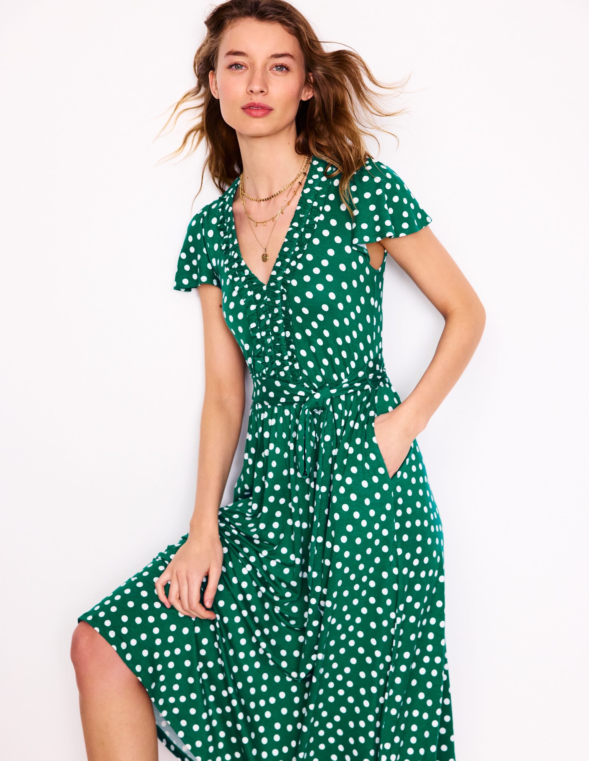 A woman wearing a green midi dress with white painted dots, ruffled neckline, and side pockets.